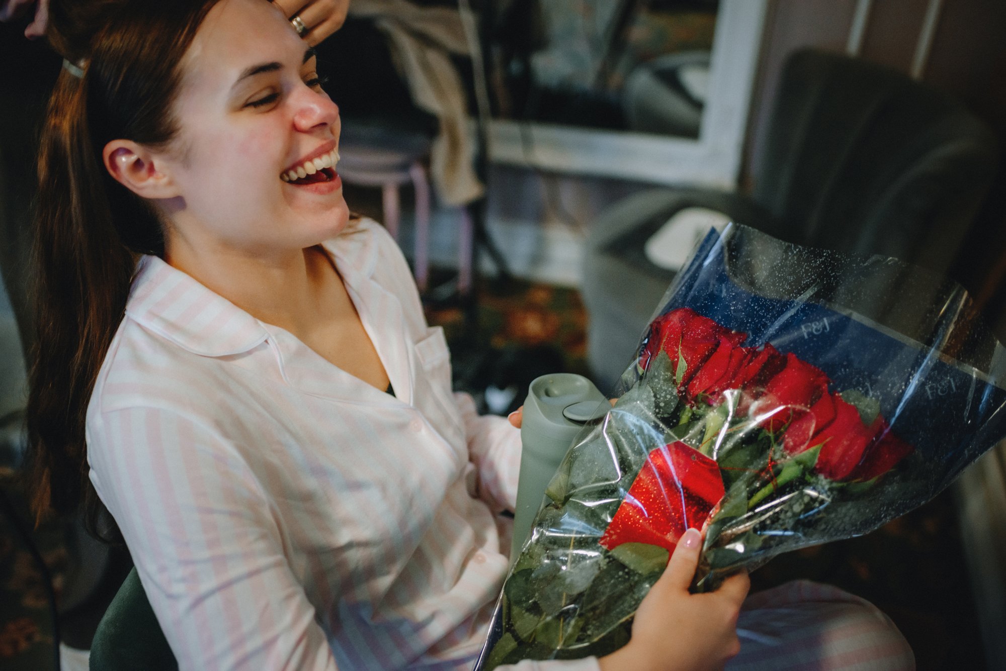 A woman in pink pajamas is smiling while holding a bouquet of red roses wrapped in clear plastic.