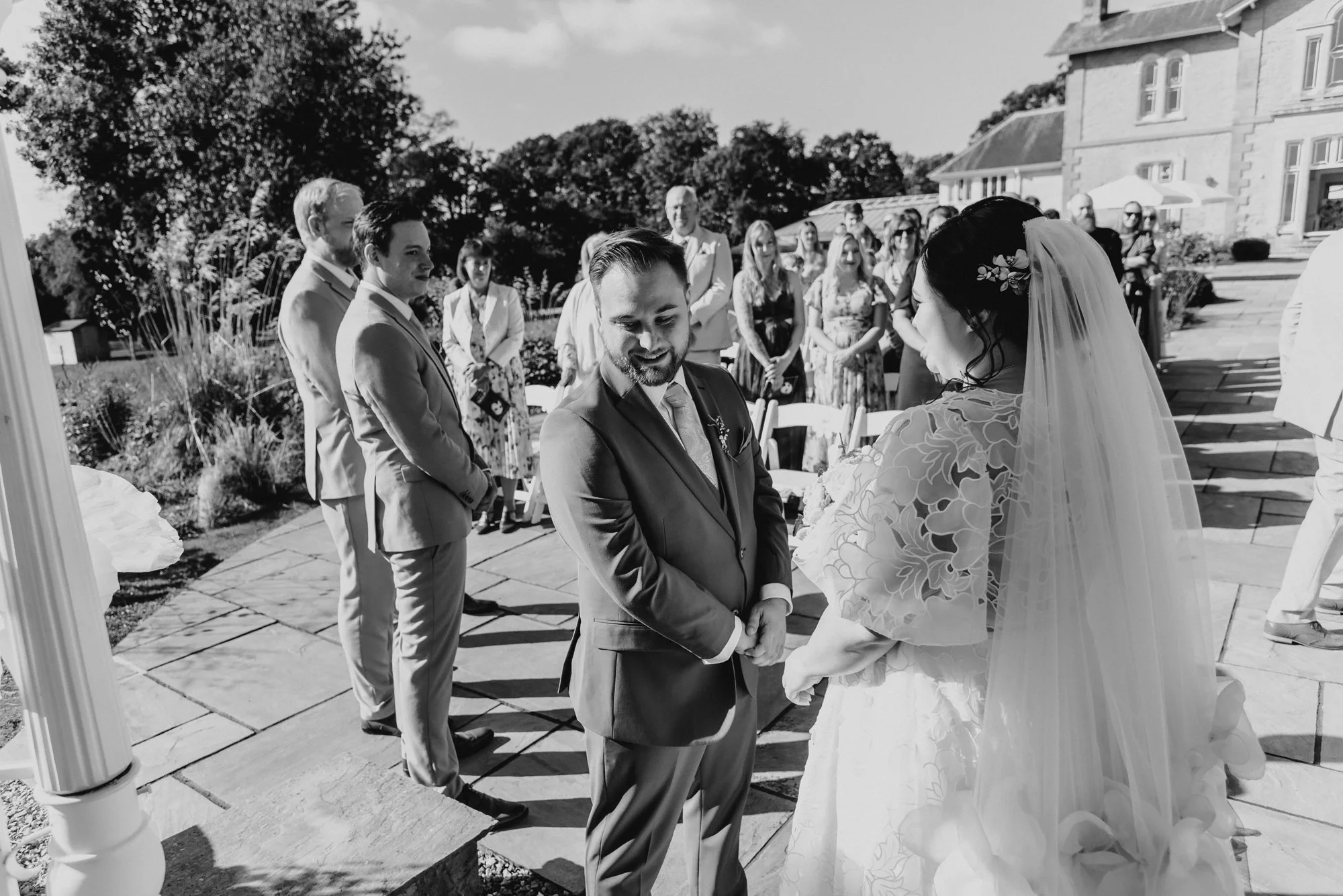 A wedding ceremony outdoors with the bride and groom exchanging vows, surrounded by guests, at a large house or estate in black and white.