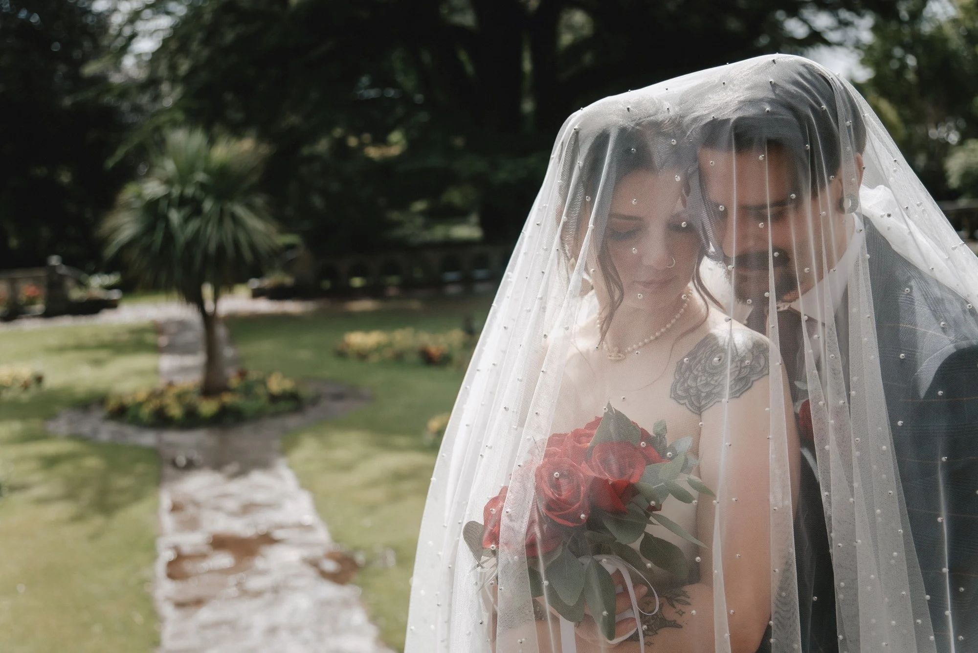 A bride and groom standing close together under a veil, with the bride holding a bouquet of red roses, in a garden setting.