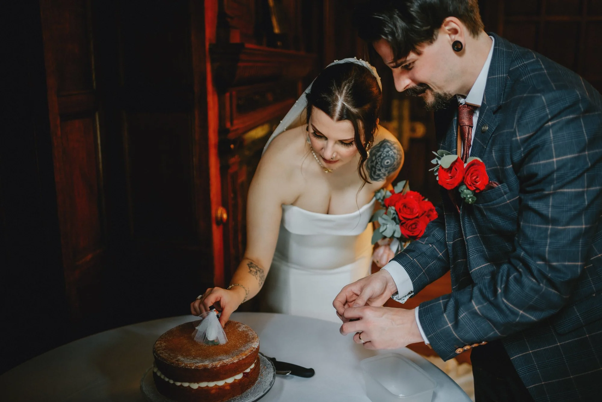 A bride and groom cutting their wedding cake. The bride is holding a cake knife and the groom is assisting. The bride has dark hair, tattoos, and is wearing a strapless white dress. The groom is wearing a checkered suit and boutonnière with red roses
