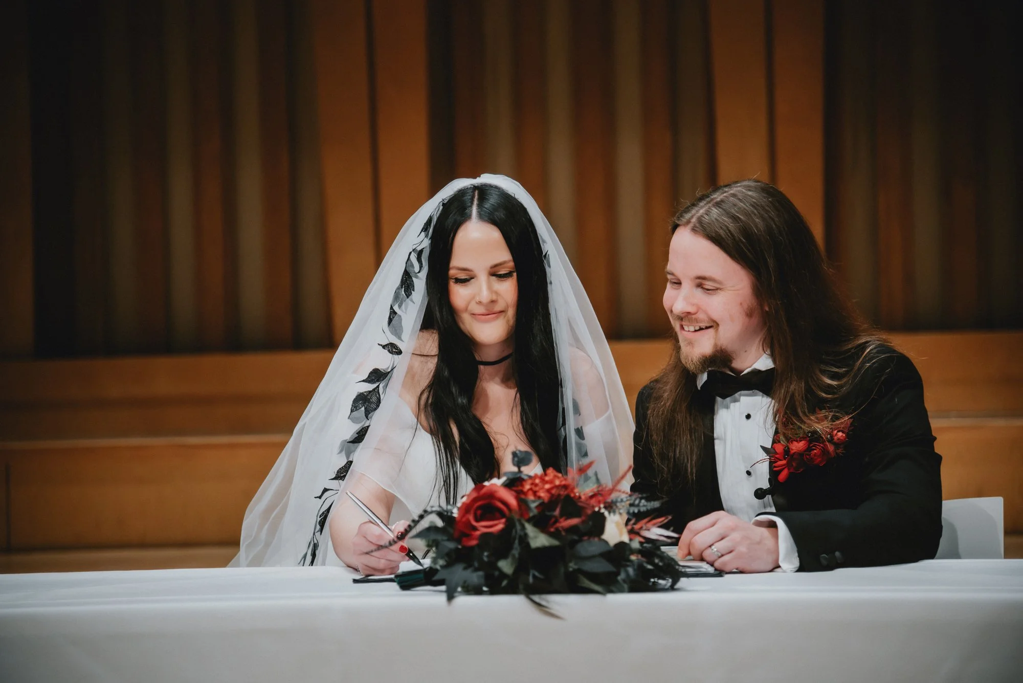 A bride with long black hair and a white veil signing a marriage register while a groom with long hair in a tuxedo with a red flower boutonniere watches and smiles during a wedding ceremony.