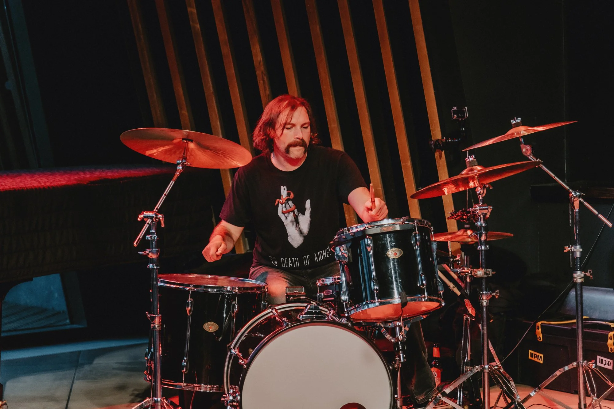 A man with long hair and a mustache playing a drum set in a music studio.