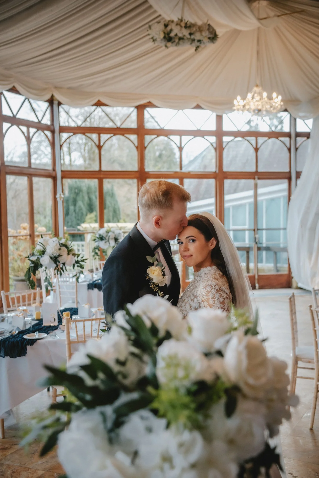 A bride and groom share a kiss at their wedding reception in a glass-enclosed venue with floral decor.