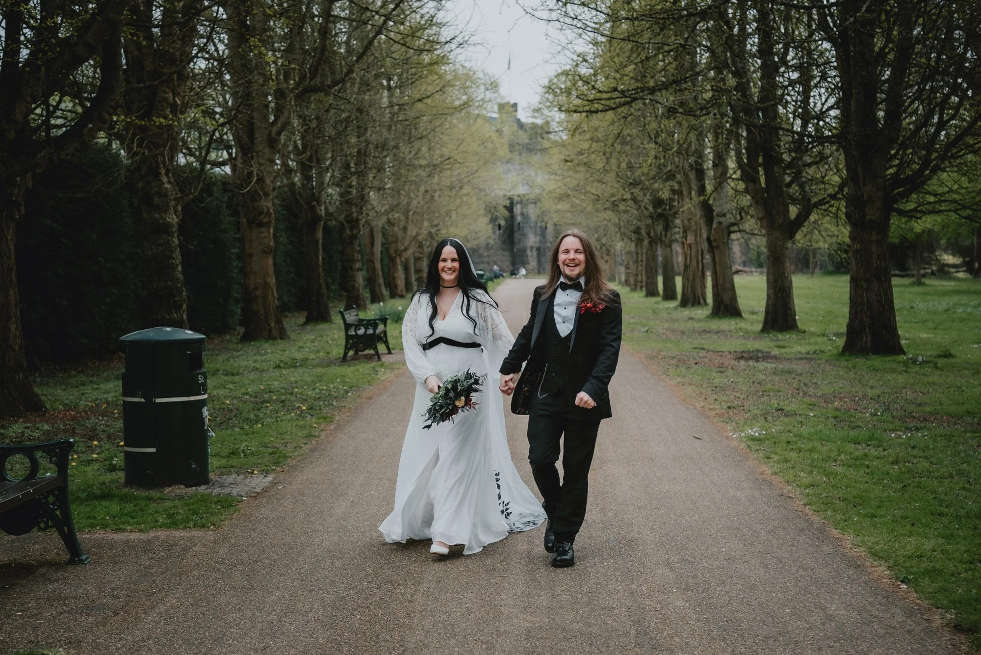 A happy couple in wedding attire walking down a park pathway holding hands, surrounded by trees and greenery.