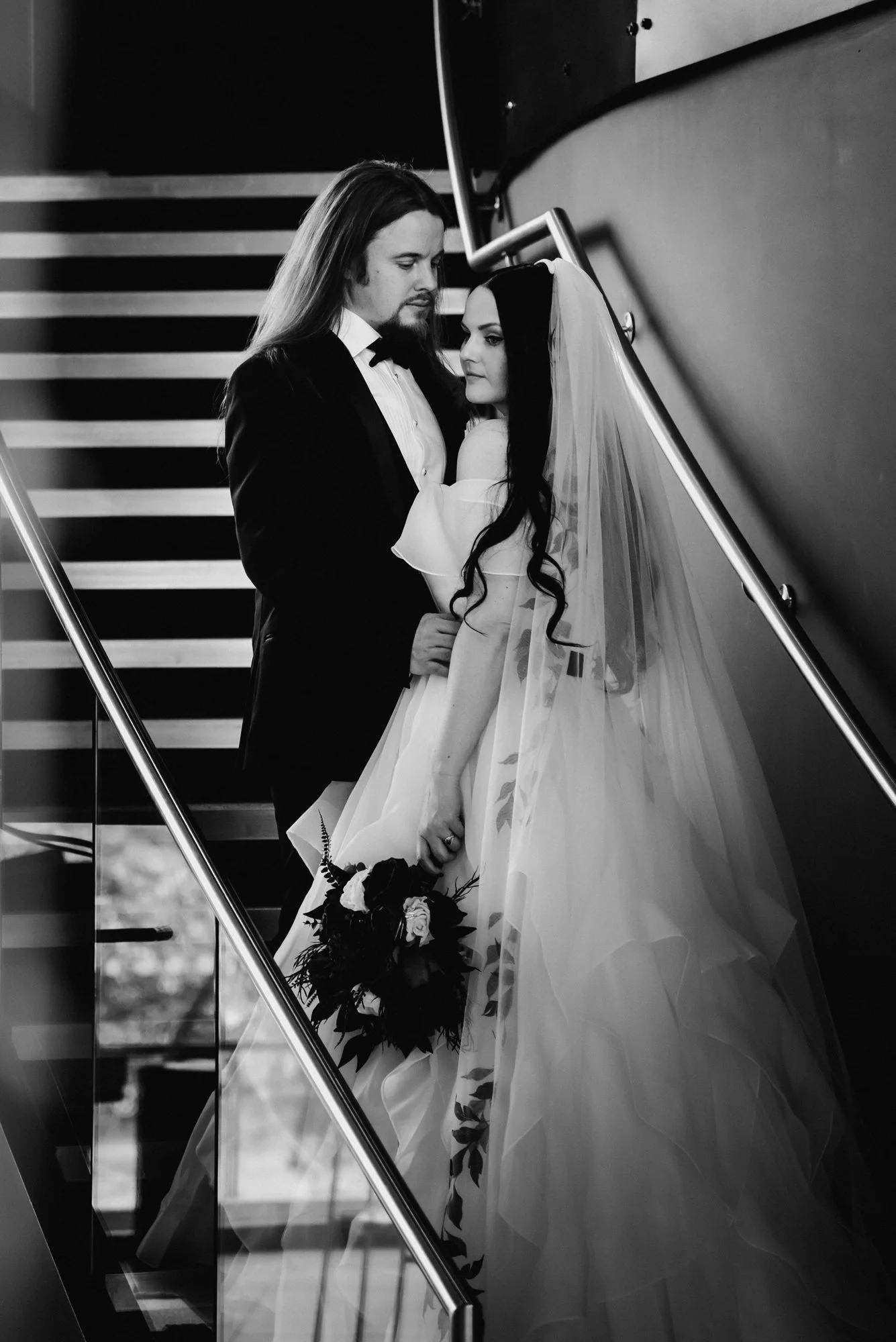 Black and white photo of a bride and groom on a staircase, gazing at each other. The bride wears a wedding gown with a veil and holds a bouquet, while the groom is in a tuxedo.