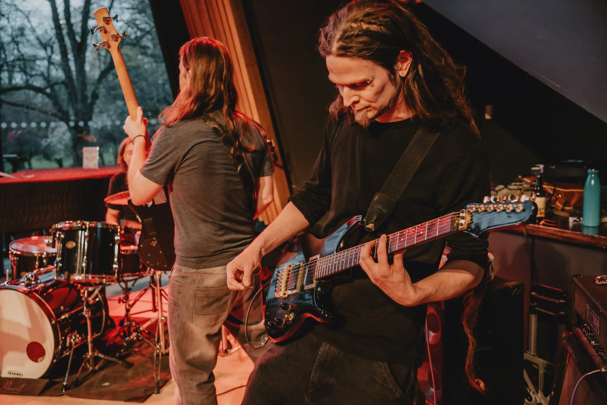 Musicians performing in a dimly lit music studio, playing guitars with drum set in the background.