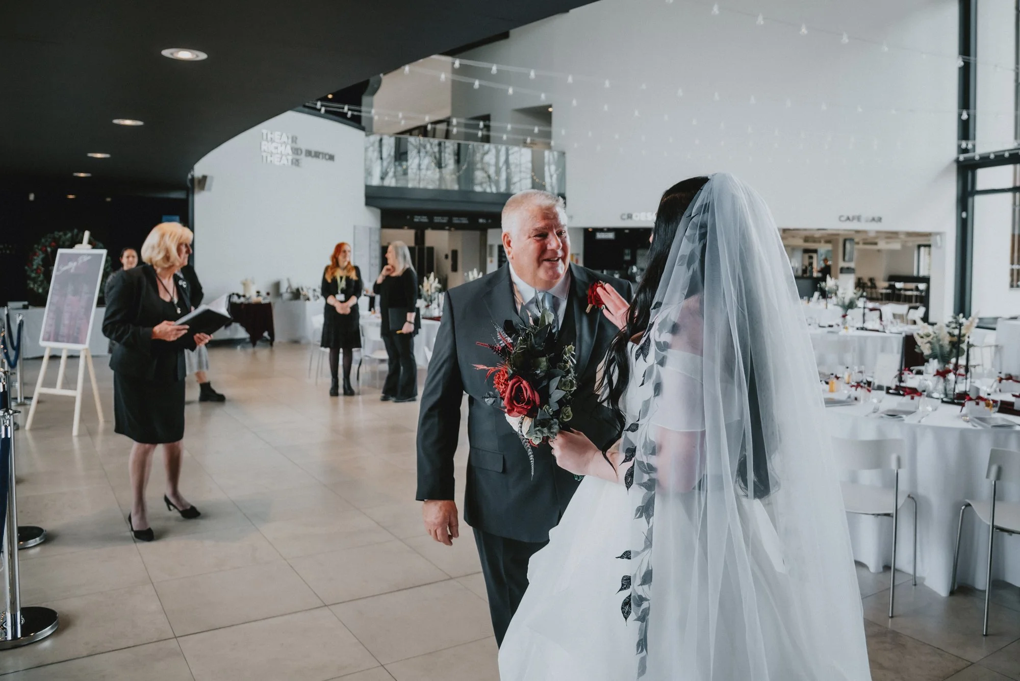 A bride in a white wedding dress and veil holding a bouquet of dark red and black flowers talking to an elderly man in a suit at a wedding reception venue. Other guests are visible in the background, along with decorated tables and string lights.