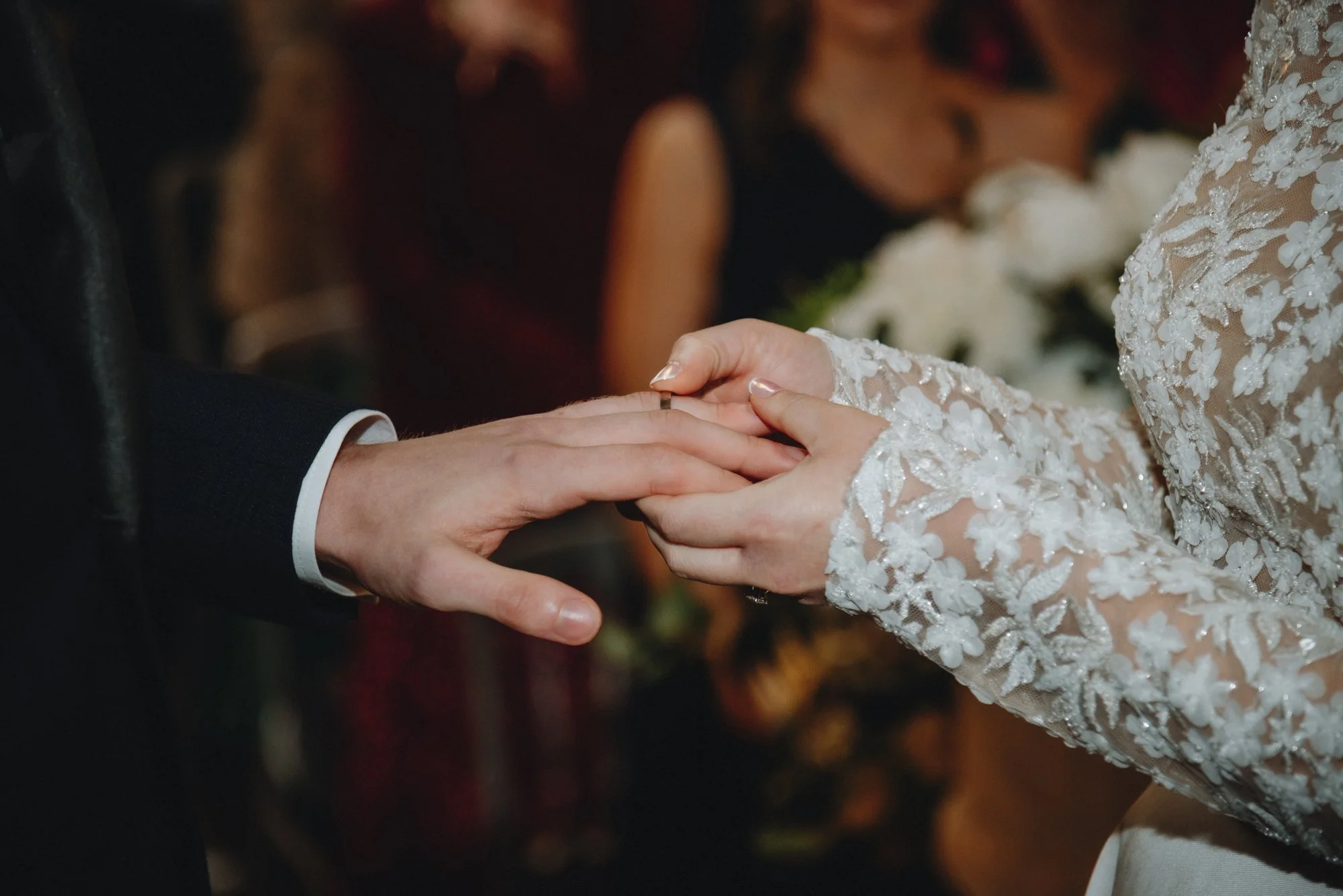 Close-up of a groom and bride holding hands during a wedding ceremony, with the groom wearing a black suit and the bride in a lace wedding dress.