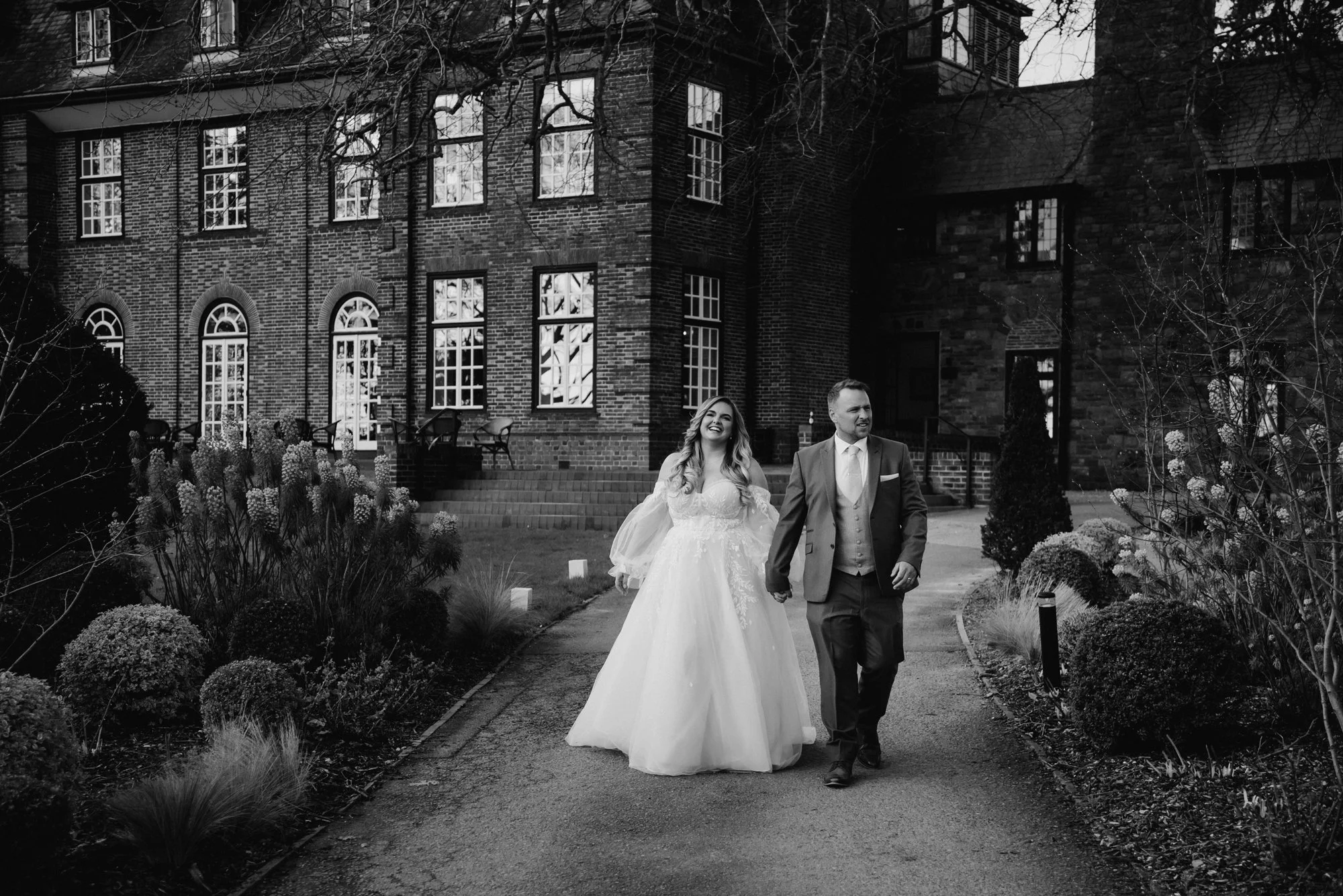 A black-and-white photo of a bride and groom holding hands and walking outside a brick building with large windows, surrounded by plants and bushes.