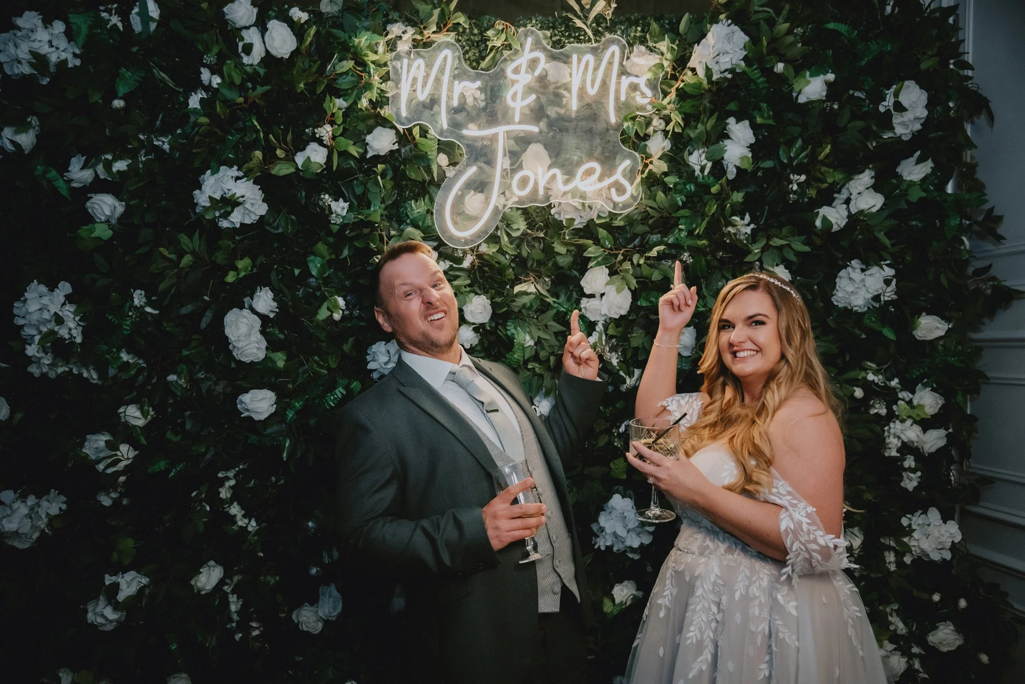 A newlywed couple standing in front of a floral backdrop with a neon sign reading "Mr & Mrs Jones," smiling and pointing at the sign while holding glasses of wine.