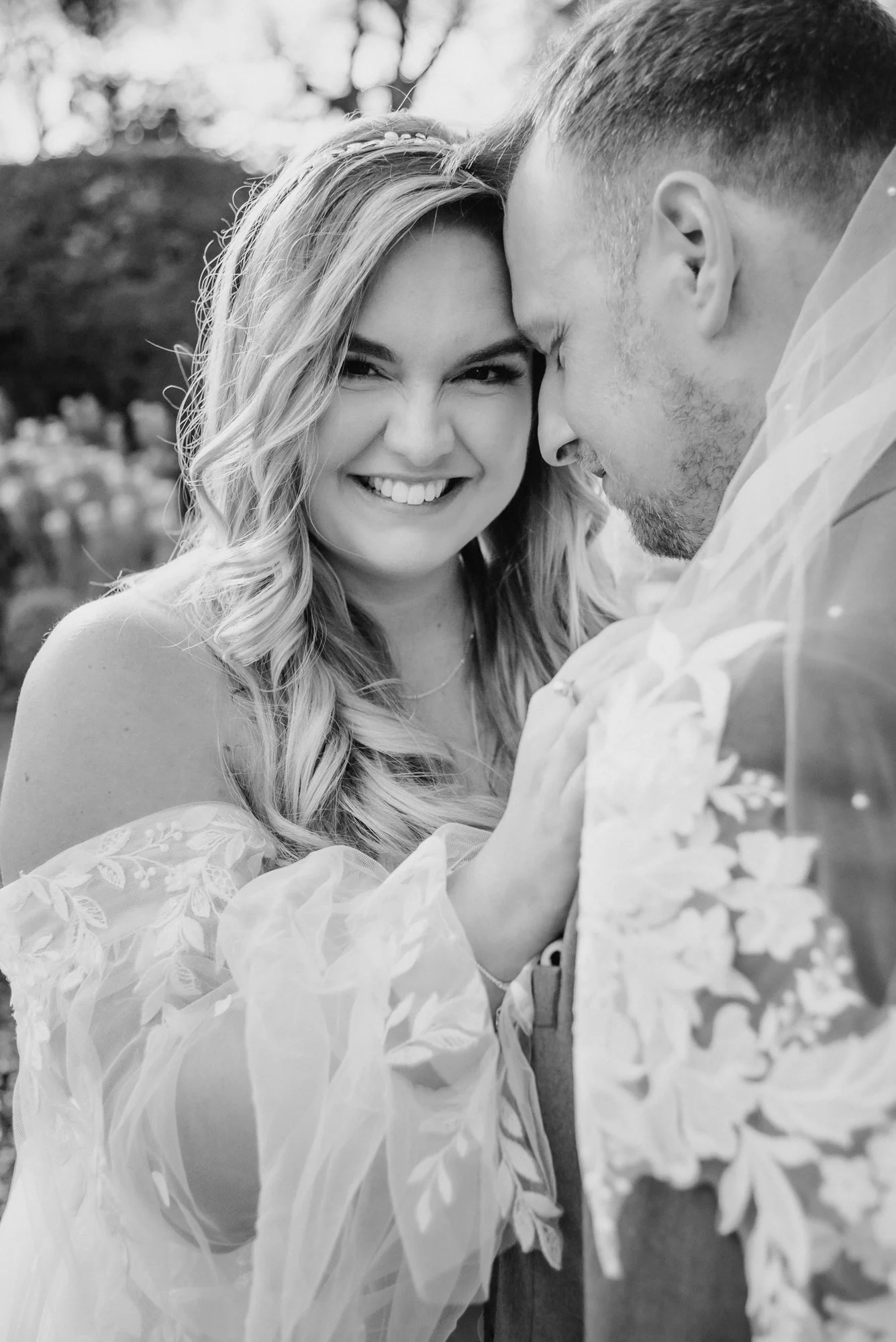 Black and white photo of a smiling woman with wavy hair and a man with short hair and facial hair, close together outdoors, with trees in the background.