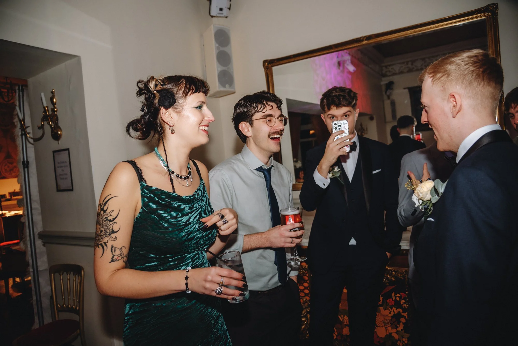 Group of five young adults dressed in formal attire enjoying socializing at an indoor event, with one person taking a photo or video on a smartphone.