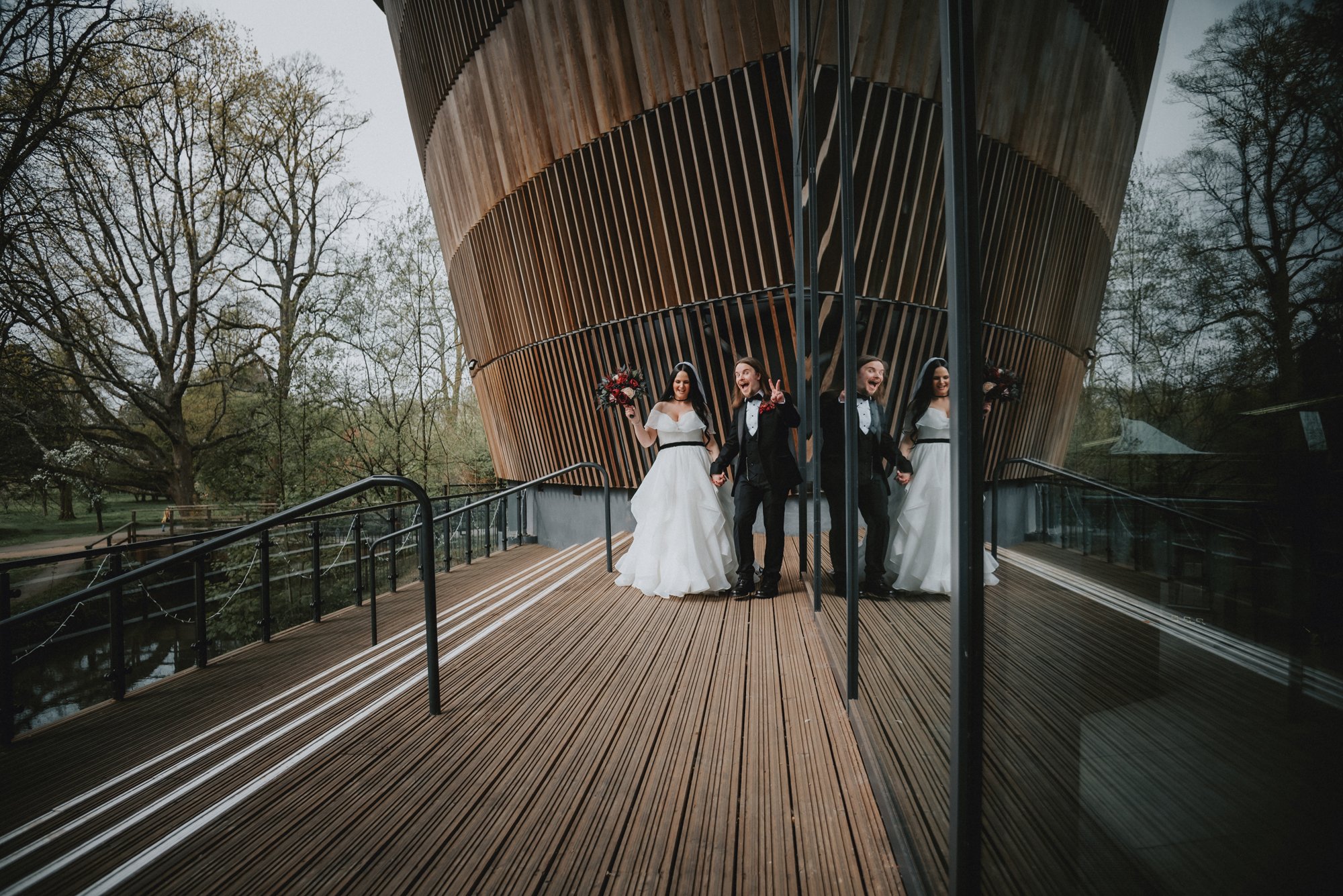 A wedding scene with two brides and a groom standing on a wooden deck with a modern, curved wooden building in the background. The brides are wearing white wedding dresses, holding flowers, and the groom is in a black tuxedo. They are reflected in a 