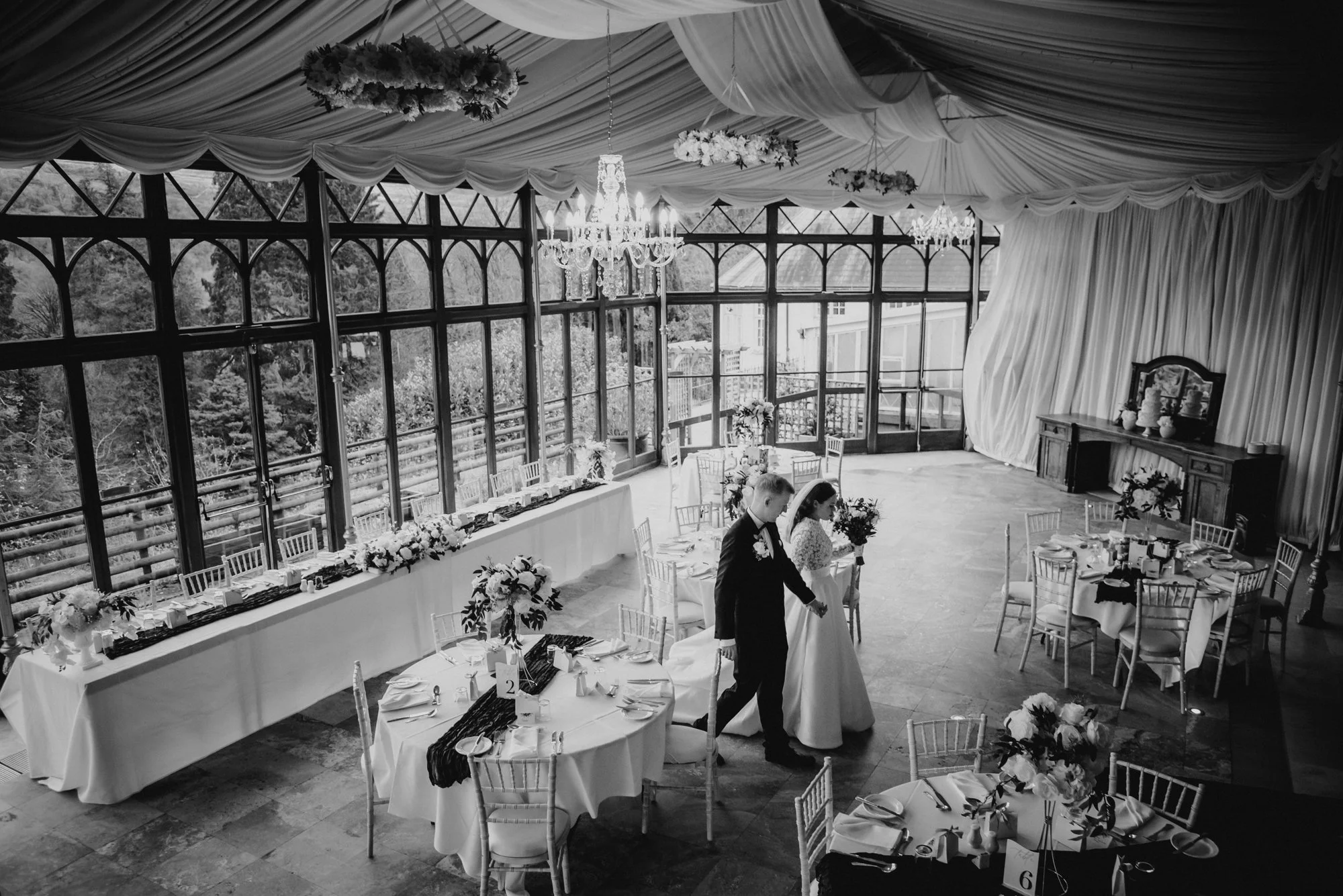 Black and white photograph of an elegant wedding reception in a glass-walled venue with a high, decorated ceiling, chandeliers, and floral arrangements on the tables. A bride and groom stand near a set table, holding hands, with other tables and chai