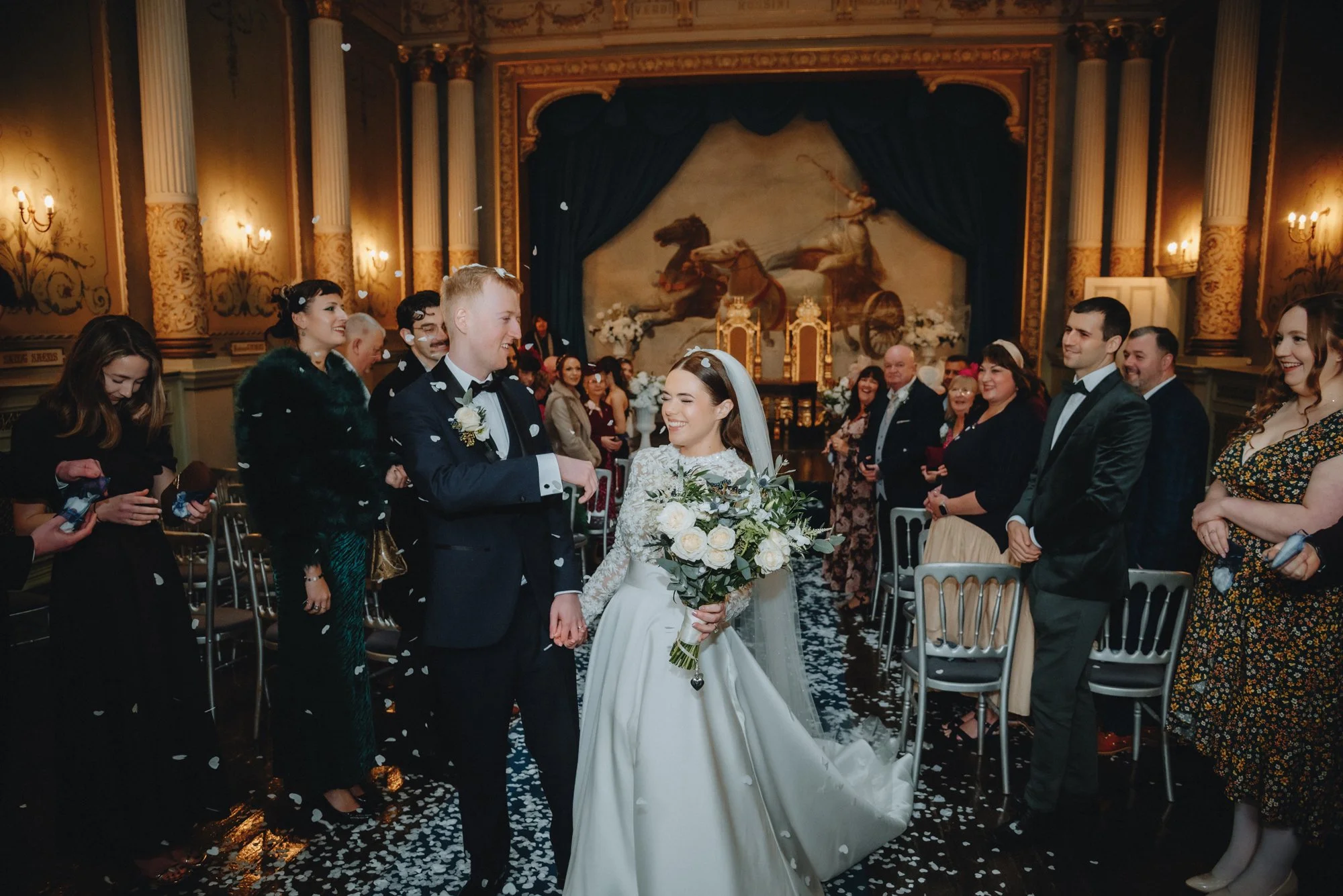 Bride and groom holding hands, smiling, with guests standing on either side, inside a decorated, elegant venue with a large painting of horses and chariots in the background, as confetti falls.