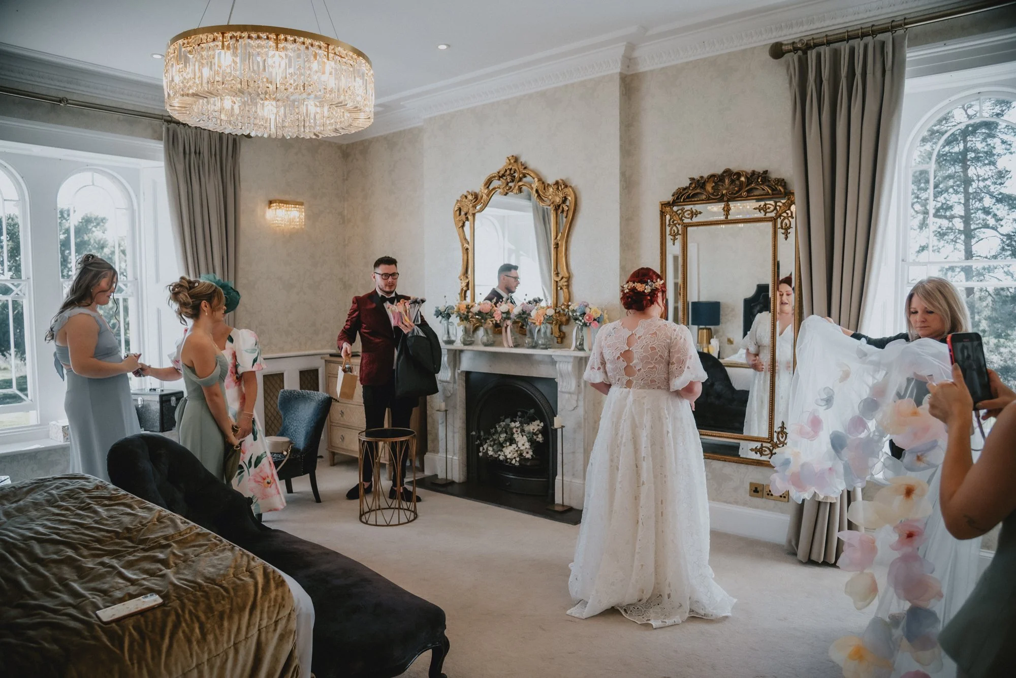 A bride in a white lace wedding dress with a red floral headpiece stands in a luxurious, well-decorated room with large windows, surrounded by women and a man. Some women are taking photos, and others are preparing or in conversation. The room featur