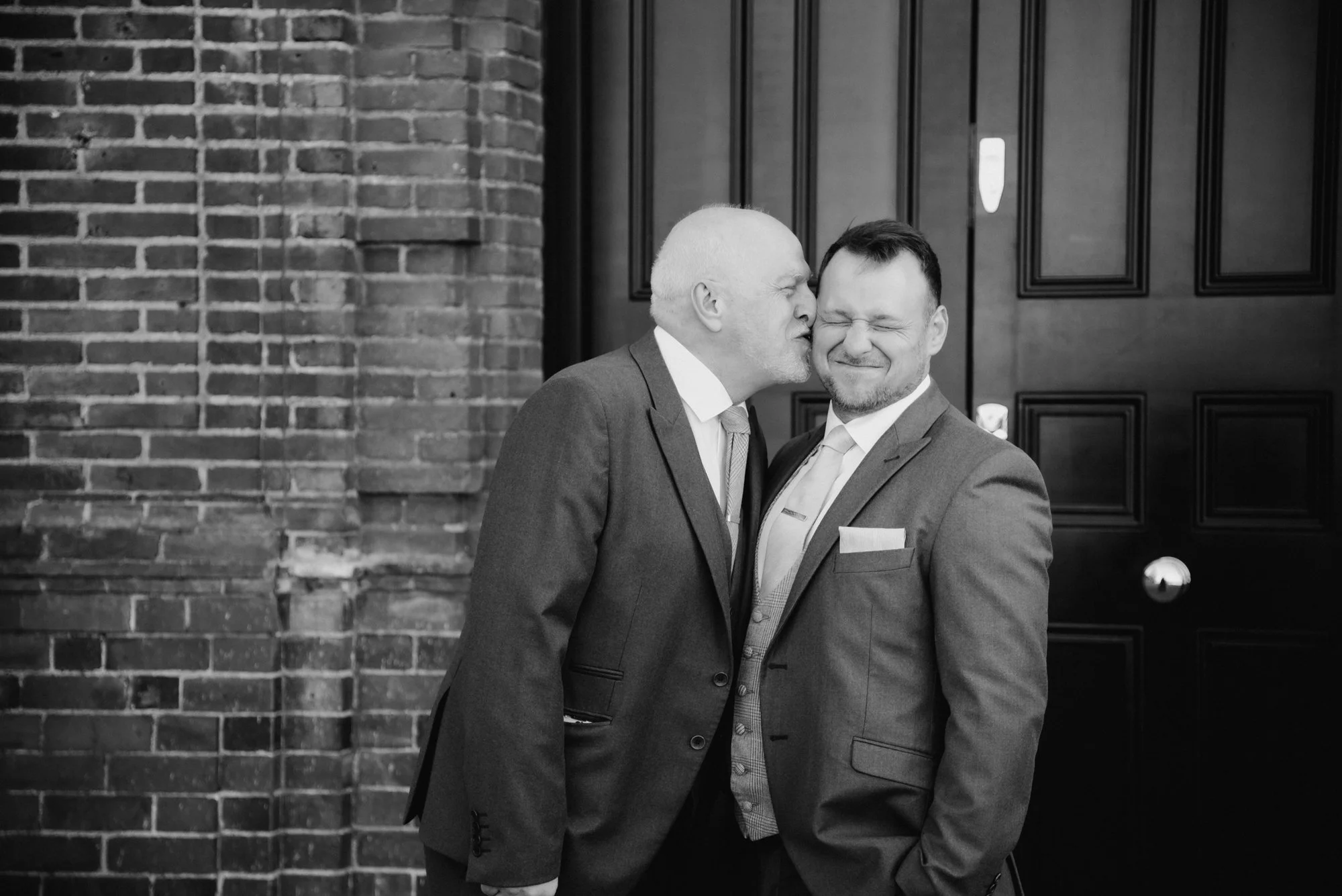 A man in a suit giving a kiss on the cheek to another man in a suit, who is smiling with eyes closed, standing in front of a brick wall and a wooden door.
