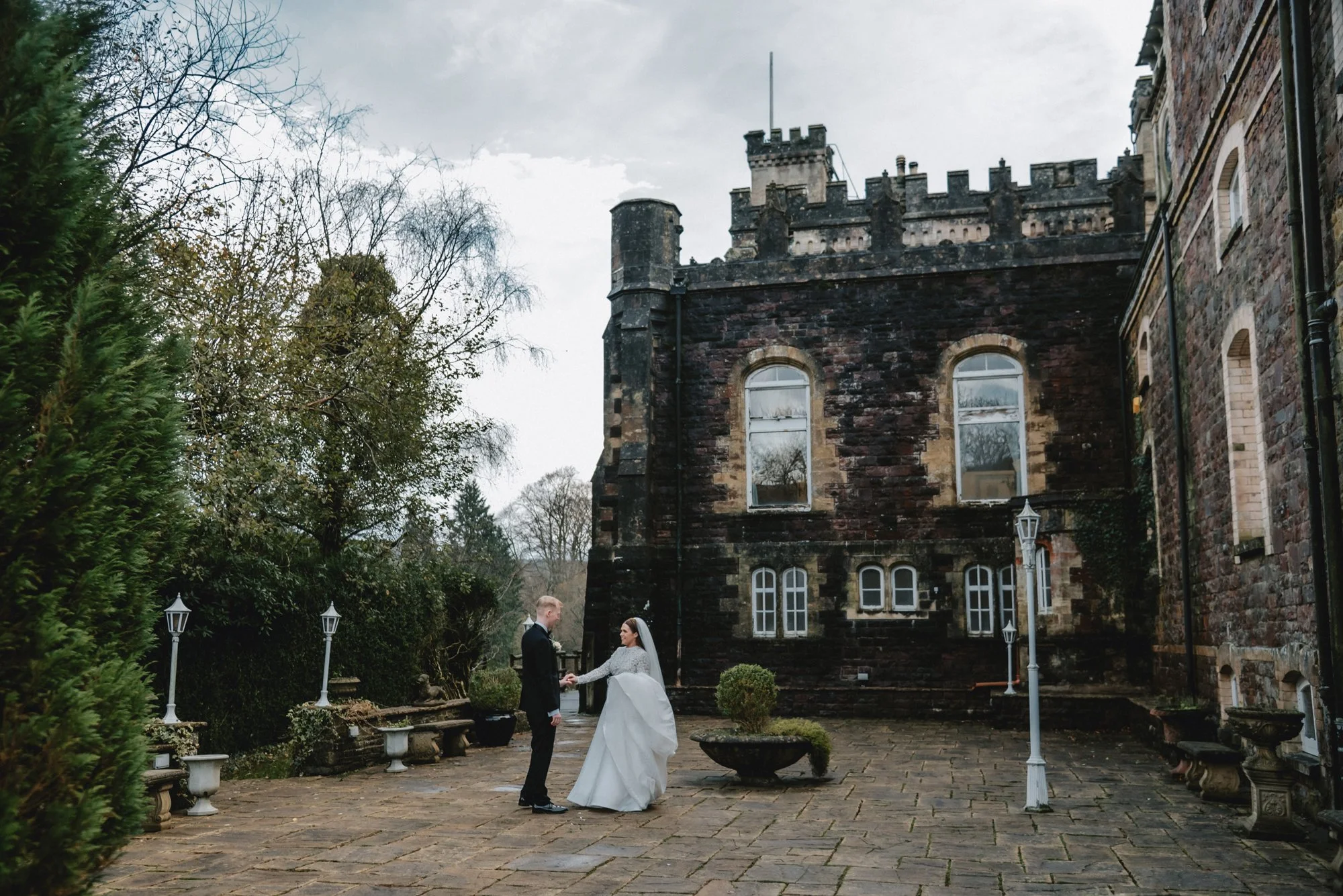 A bride and groom holding hands and looking at each other on a stone courtyard outside an old castle or mansion, surrounded by trees and decorative lamp posts.