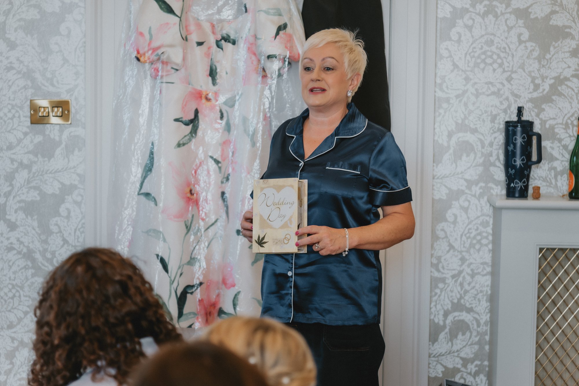 A woman with short blonde hair wearing navy satin pajamas stands inside a decorated room, holding a wedding day card. She appears to be speaking to a group of people, with their heads visible at the bottom of the image. The background includes floral