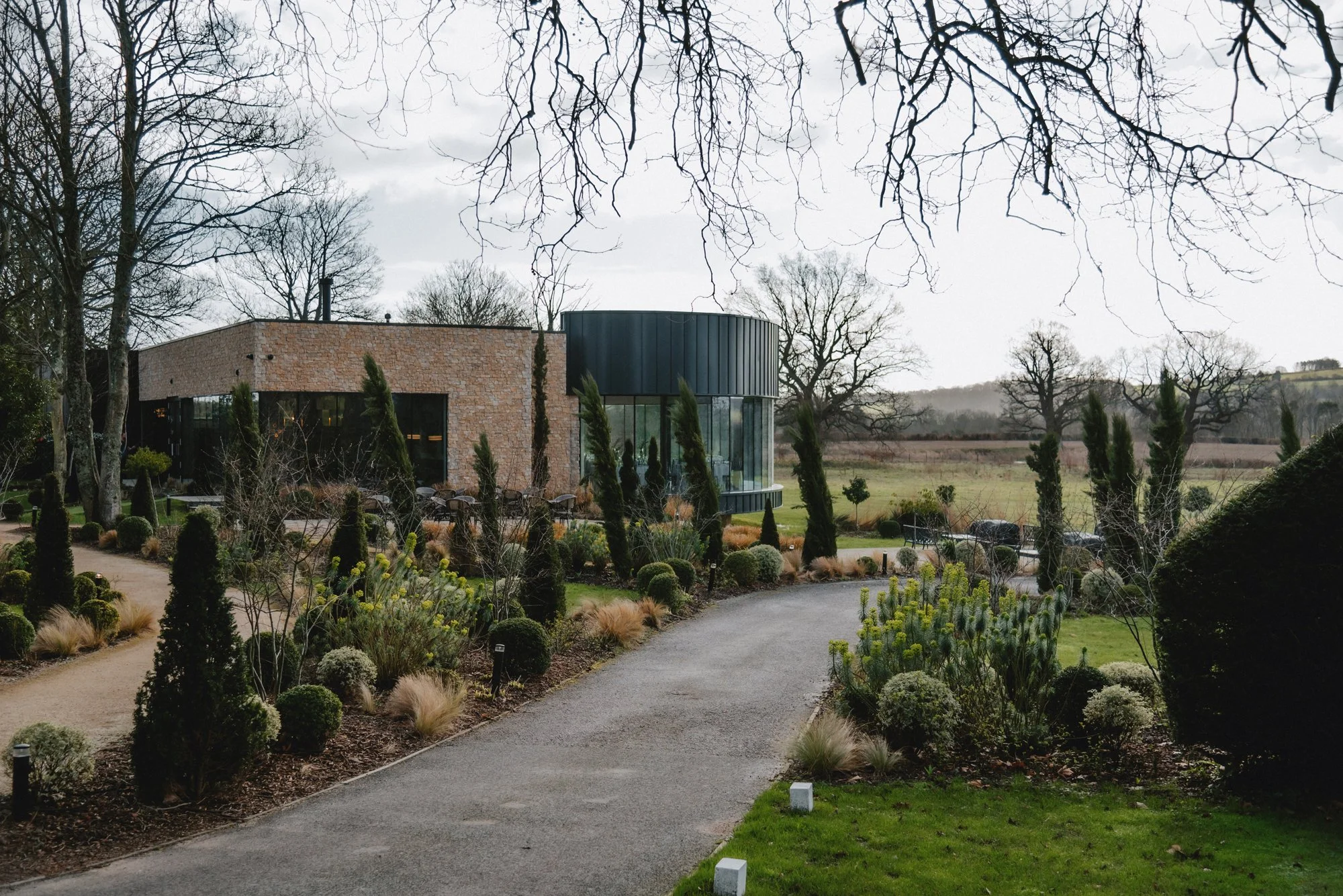 A modern building with a rounded glass facade and a brick wall, surrounded by landscaped garden with various bushes and trees, on a cloudy day.