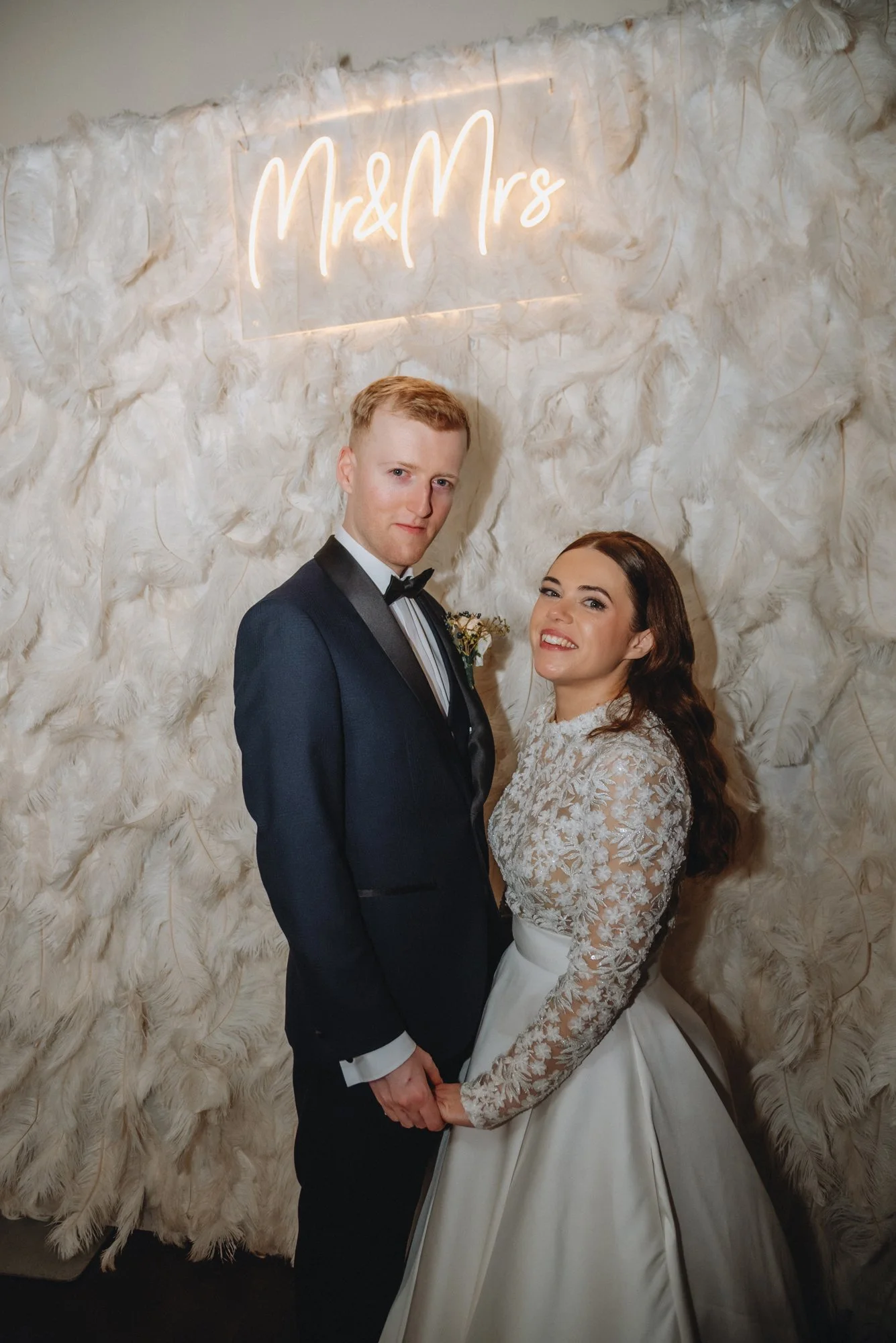 A bride and groom standing together at their wedding, holding hands, with a white feathered backdrop and a neon sign that says 'Mr & Mrs' behind them.