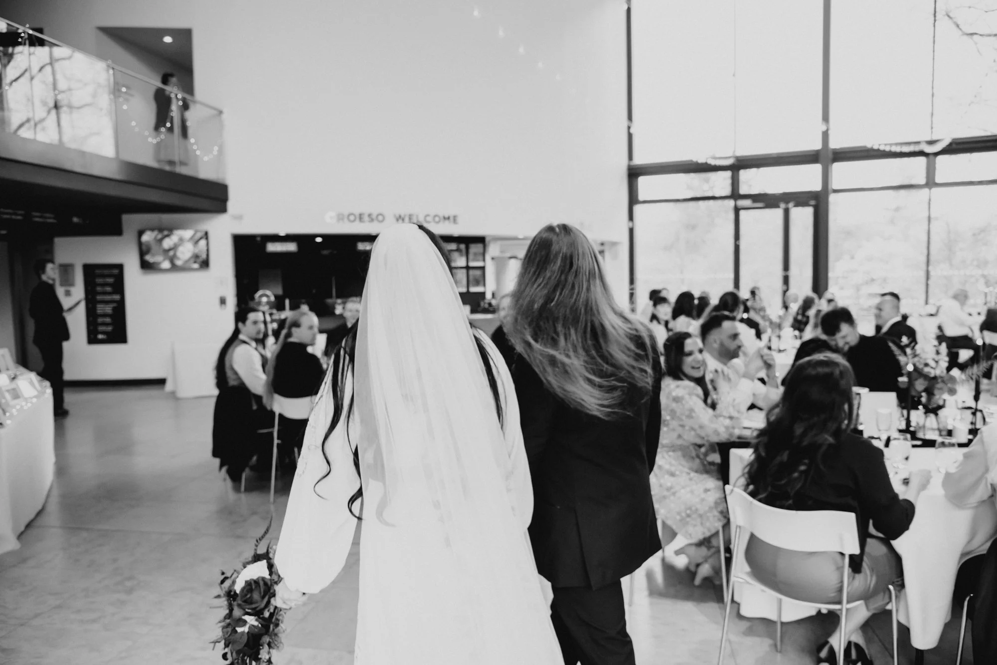 Black and white photo of a wedding reception, with the bride in a white gown and veil holding flowers, walking past guests seated at tables.