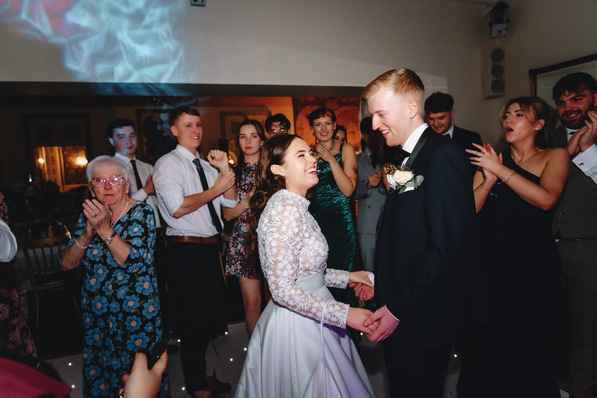 A bride and groom hold hands and smile at each other at their wedding reception, surrounded by guests clapping and celebrating indoors.
