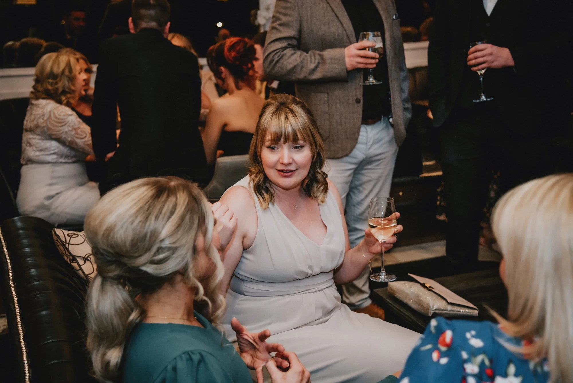 People socializing at a party, including a woman in a white dress holding a glass of wine, engaged in conversation with other guests.