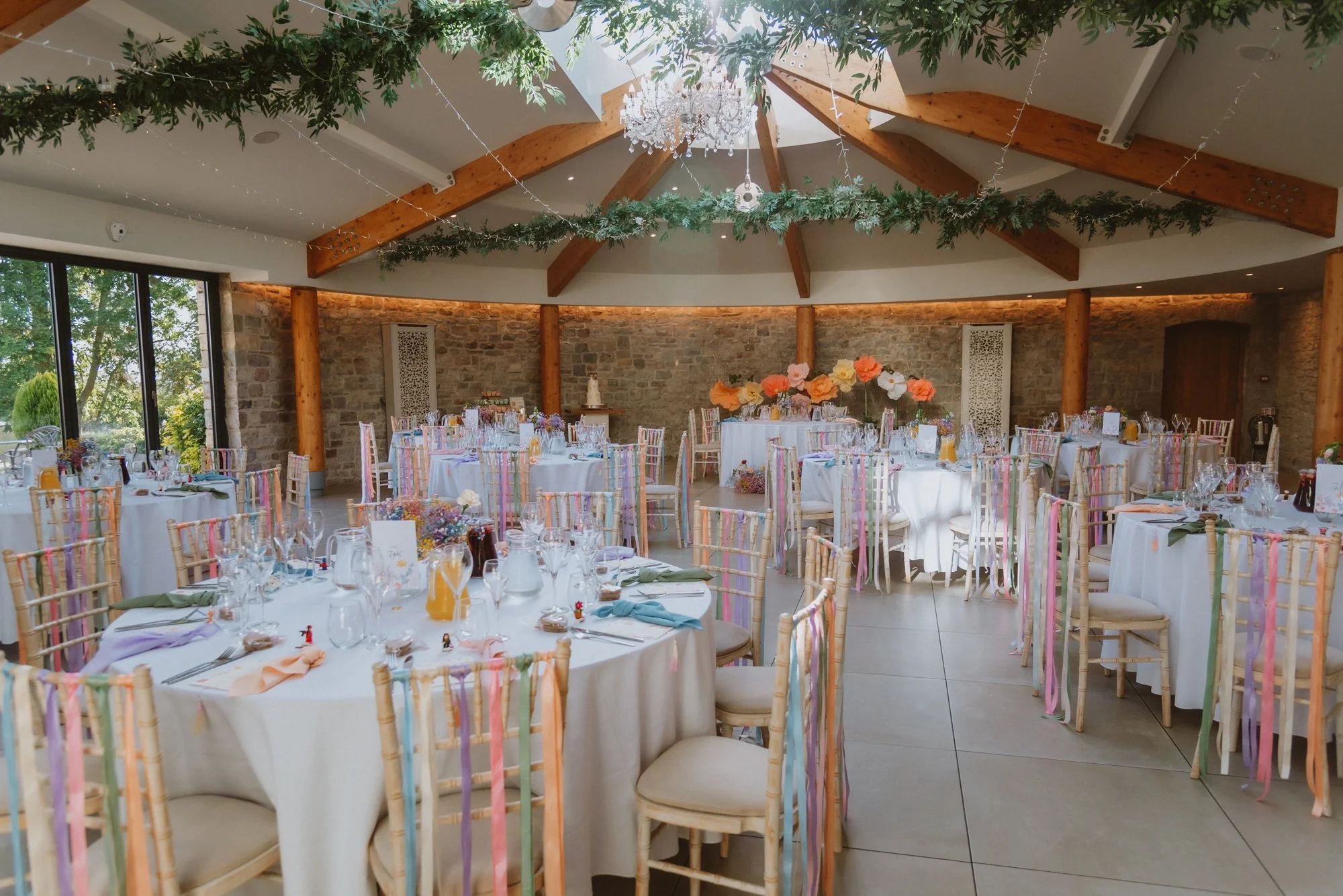 Elegant banquet hall decorated for a special event with round tables covered in white tablecloths, colorful chairs striped with pastel ribbons, floral centerpieces, and a ceiling adorned with greenery and a chandelier.