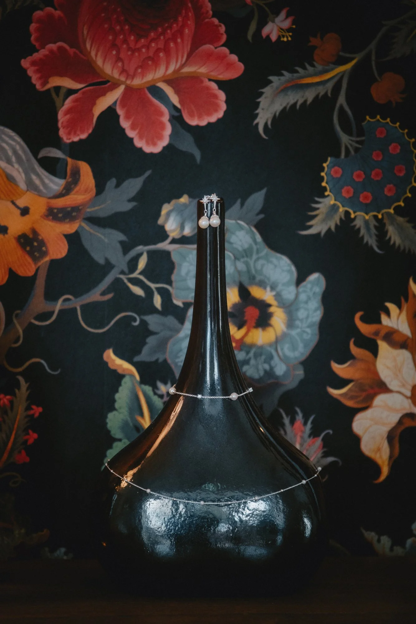 A black vase with a tall, narrow neck and round base, decorated with a string of pearls and a pearl bracelet, set against a dark floral wallpaper background.
