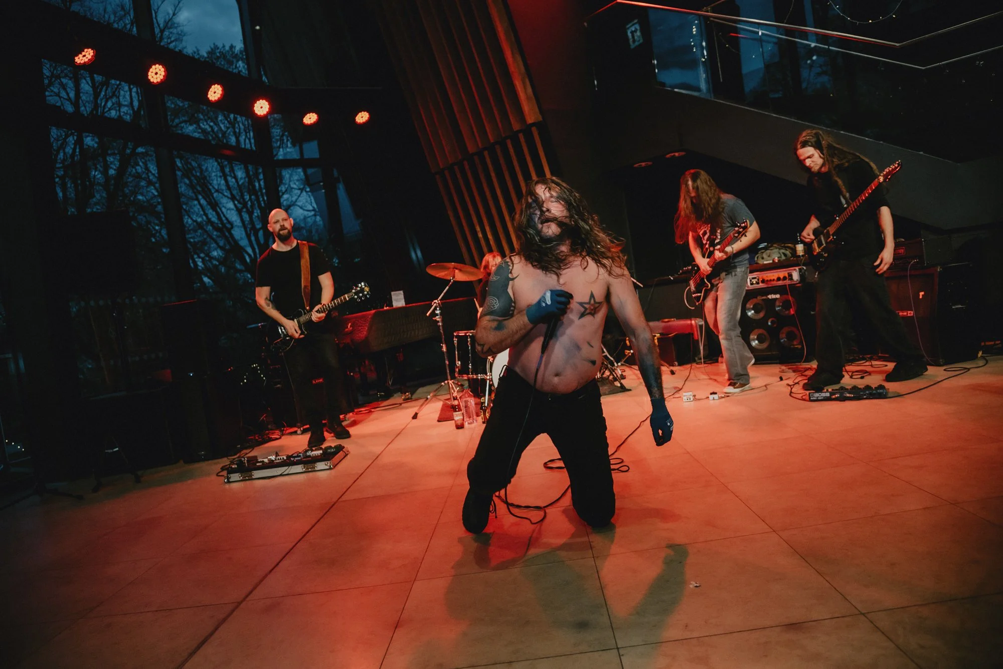 A shirtless singer with long hair, tattoos, and gloves kneels on stage holding a microphone during a rock concert, with a band playing guitars behind him in a dimly lit venue.