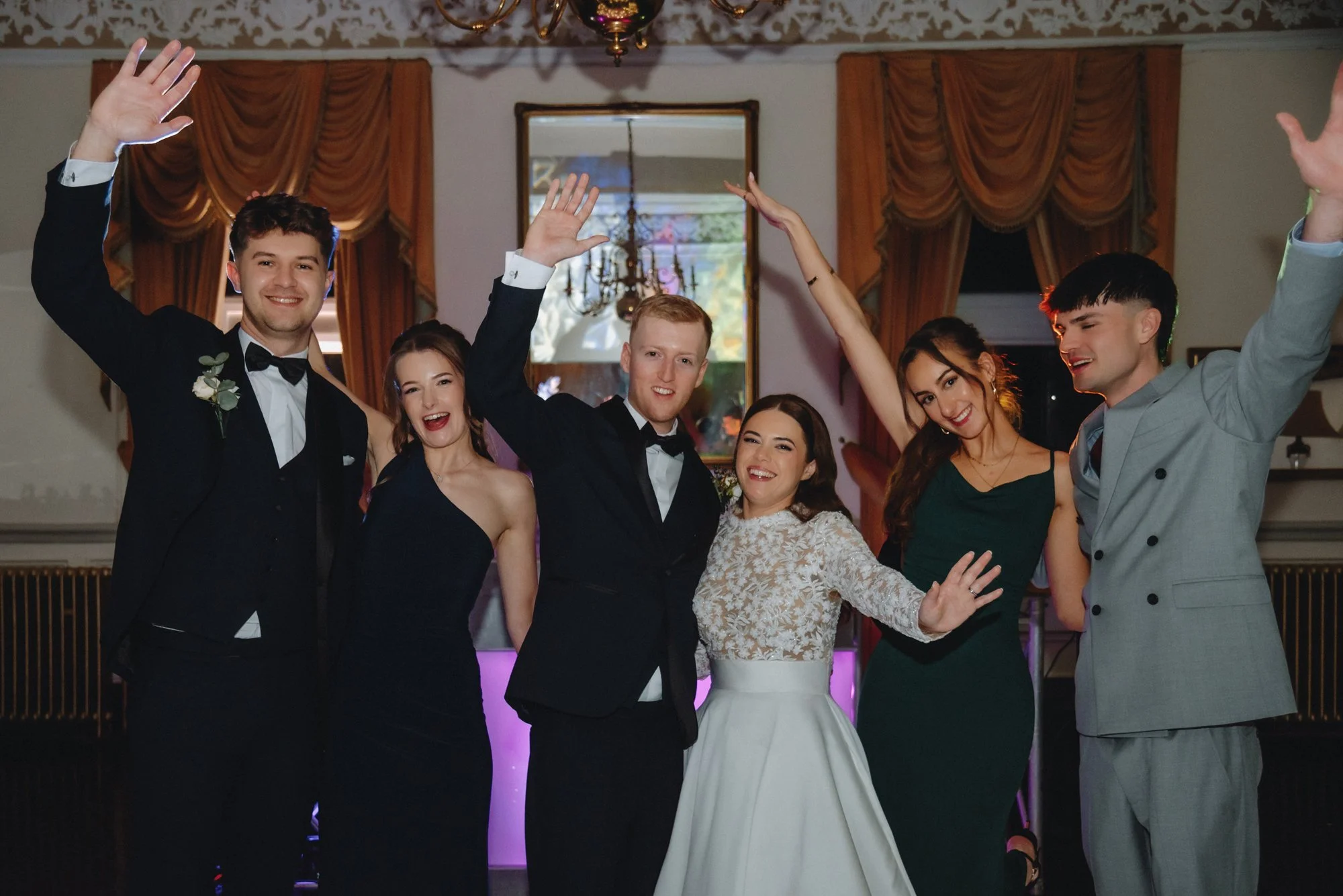 A group of six people dressed in formal attire, celebrating at a party with smiles and raised hands indoors.