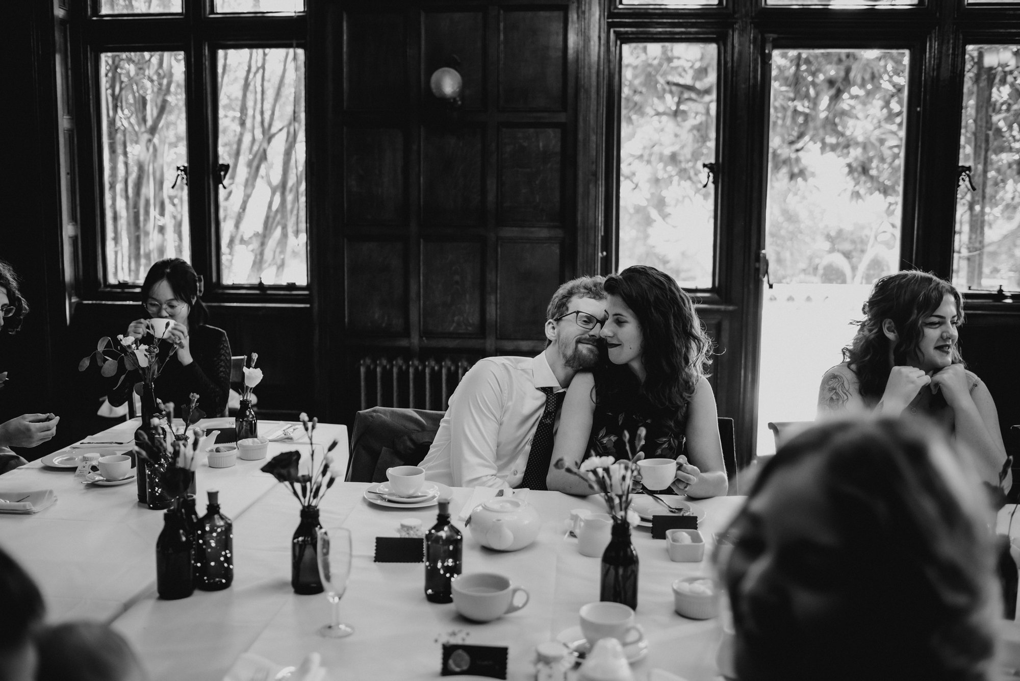 People sitting at a table during a gathering, with one couple in focus sharing a tender moment as the woman whispers to the man, both smiling. The scene is in a room with large windows and wooden paneling, with other guests in the background and tabl