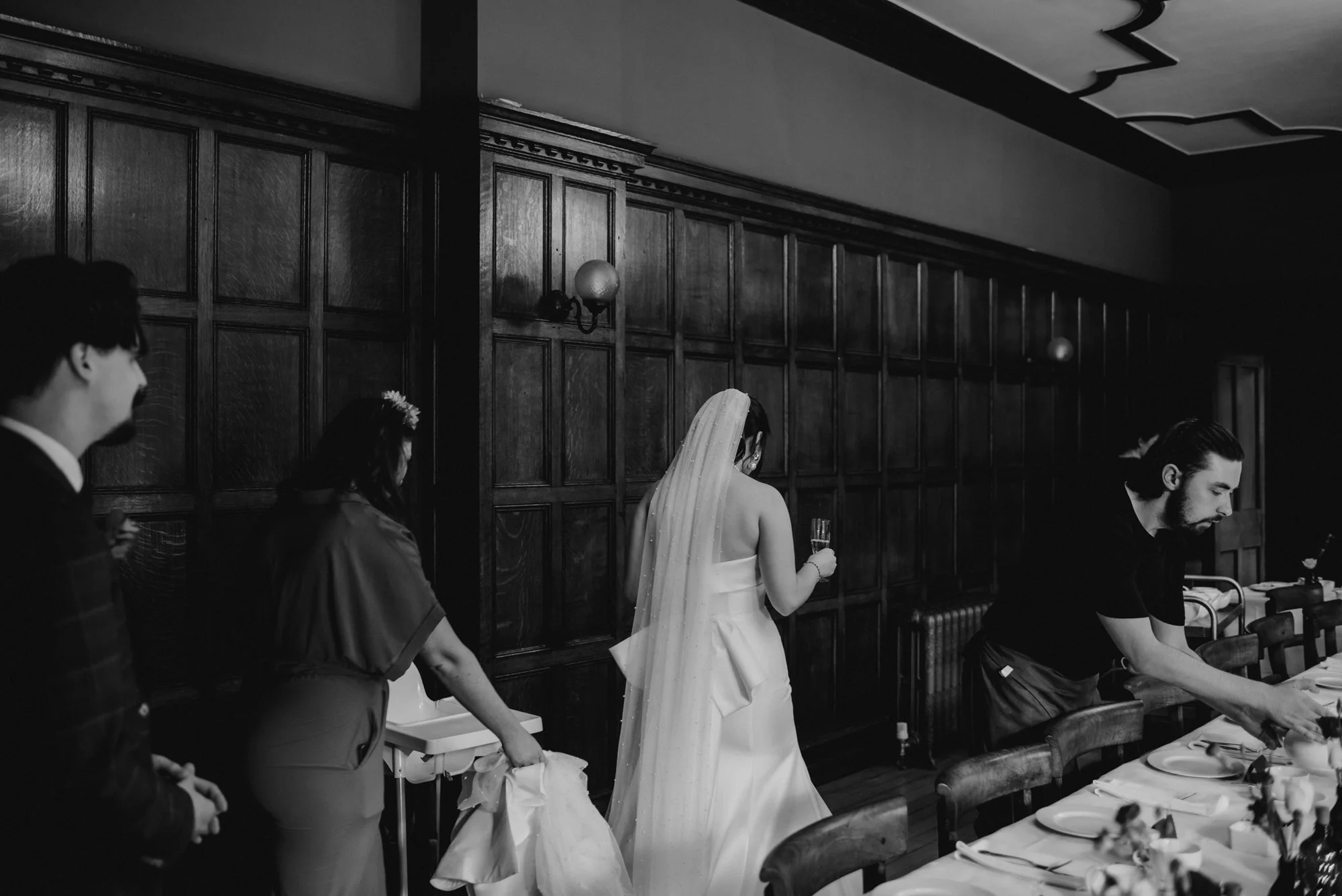 Black and white photo of a bride holding a glass of champagne in a banquet hall. She is wearing a wedding dress and veil, with her back to the camera. Other people are preparing a table for a meal, with chairs, plates, and silverware visible.