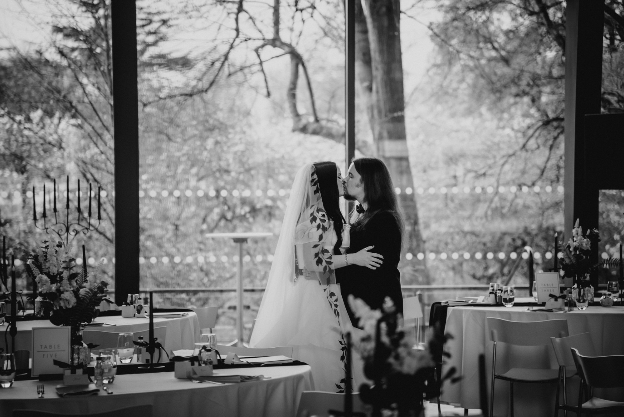 A black and white photo of two women, one in a wedding dress and veil, and the other in dark clothing, kissing each other inside a decorated event space with tables set for a celebration, large windows revealing trees outside.