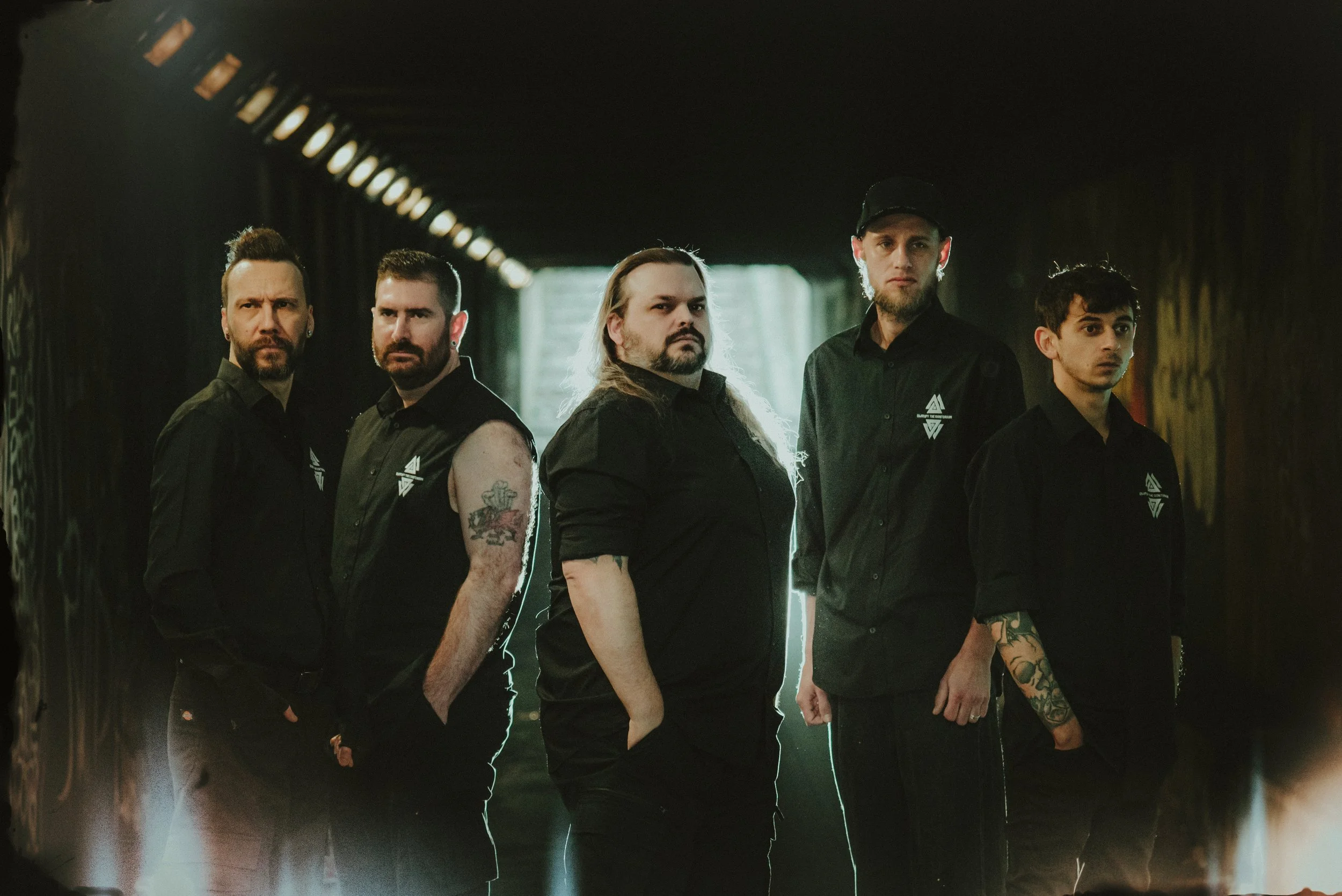 Five men standing in a dark tunnel with graffiti, dressed in black shirts, some with tattoos, looking serious.