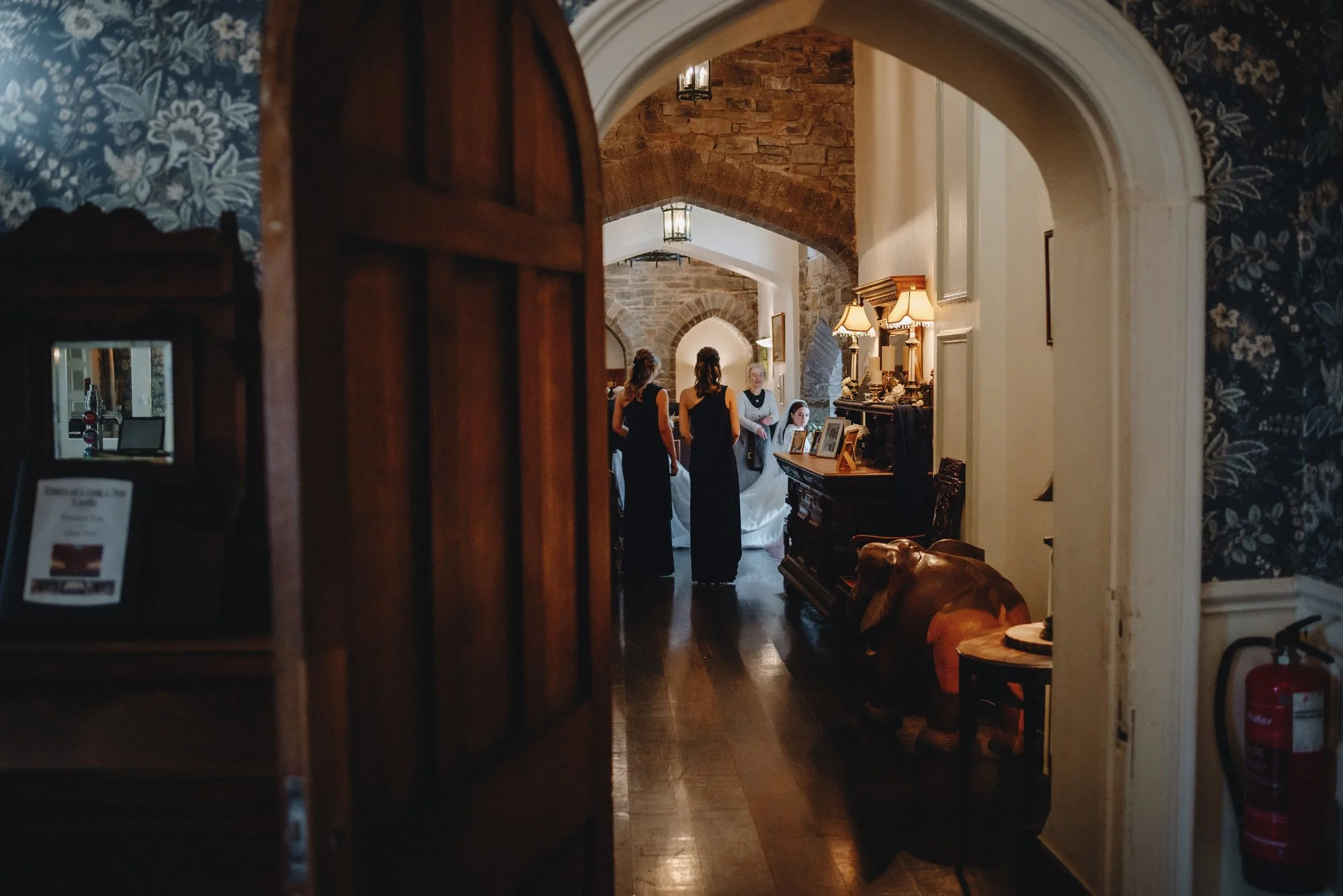 A glimpse into a cozy, vintage-style room with warm lighting, showing four women in dark dresses gathered near a fireplace, some looking at a phone or a picture, with old-fashioned lamps and photographs on the mantle, and wooden furniture in a rustic
