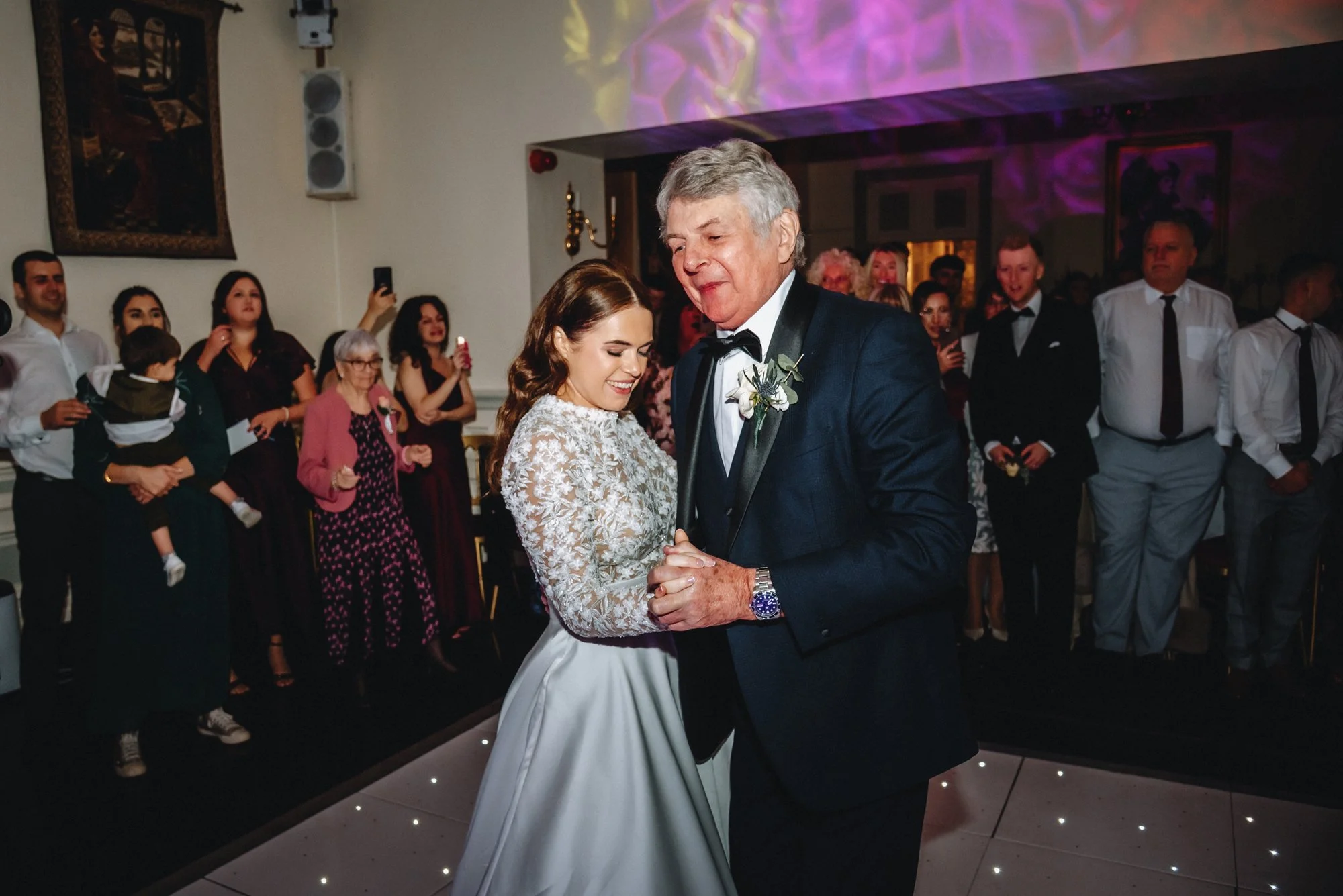 A bride and an older man, possibly her father, are dancing together at a wedding reception. The bride is wearing a white lace wedding dress, and the man is in a dark suit with a boutonnière. Guests are standing around, watching and taking photos, in 