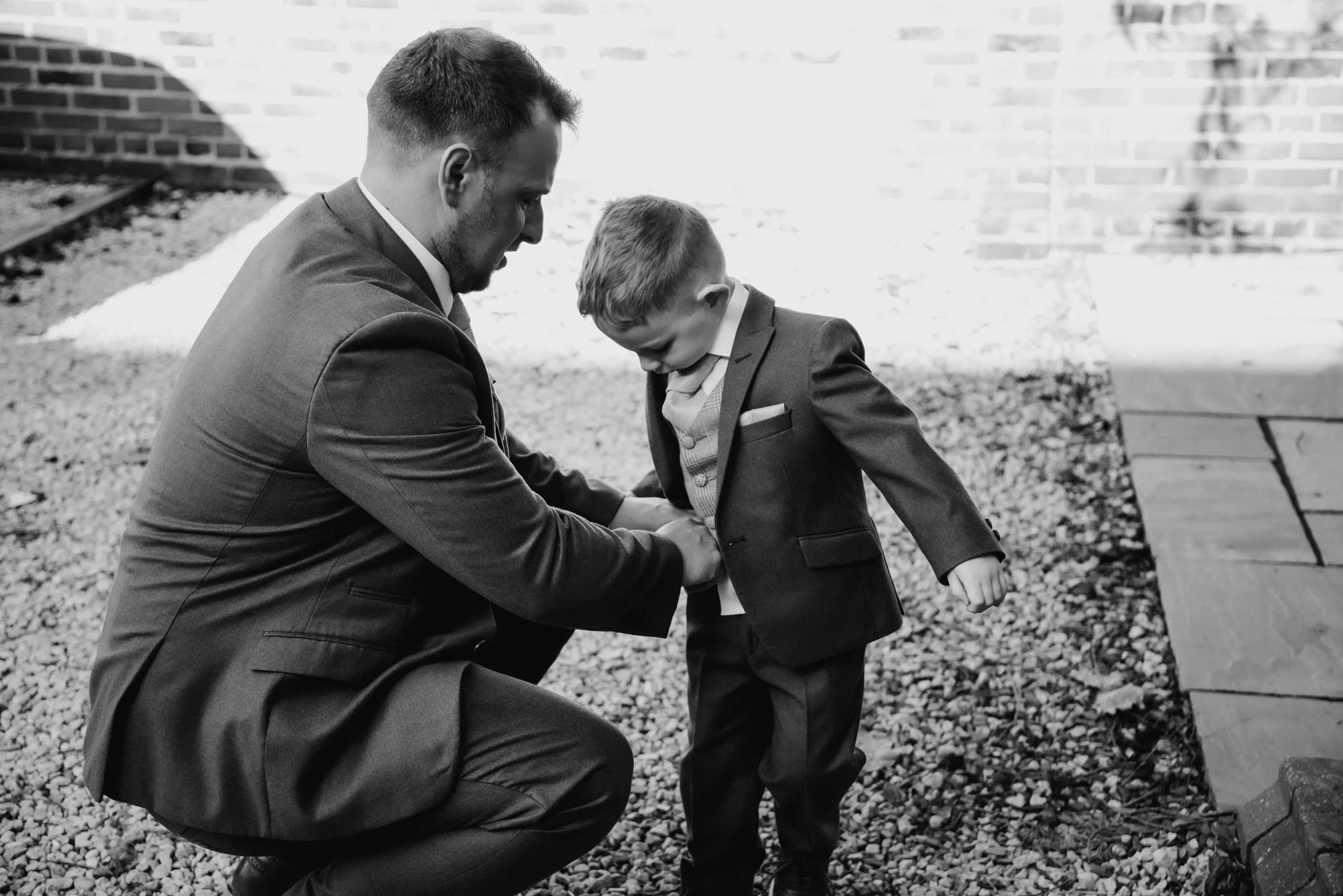 A man in a suit kneeling down and helping a young boy in a suit adjust his jacket outside on a gravel surface.
