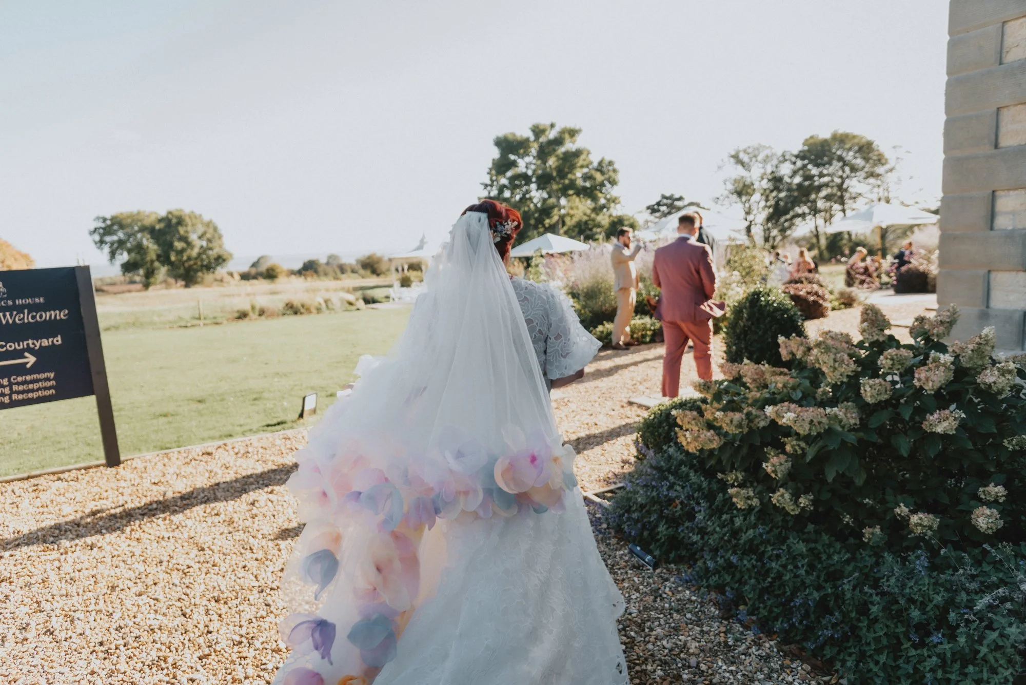 A bride with a lace dress and a long veil decorated with pastel flower petals walking outside toward a reception area, with guests and umbrellas visible in the background.