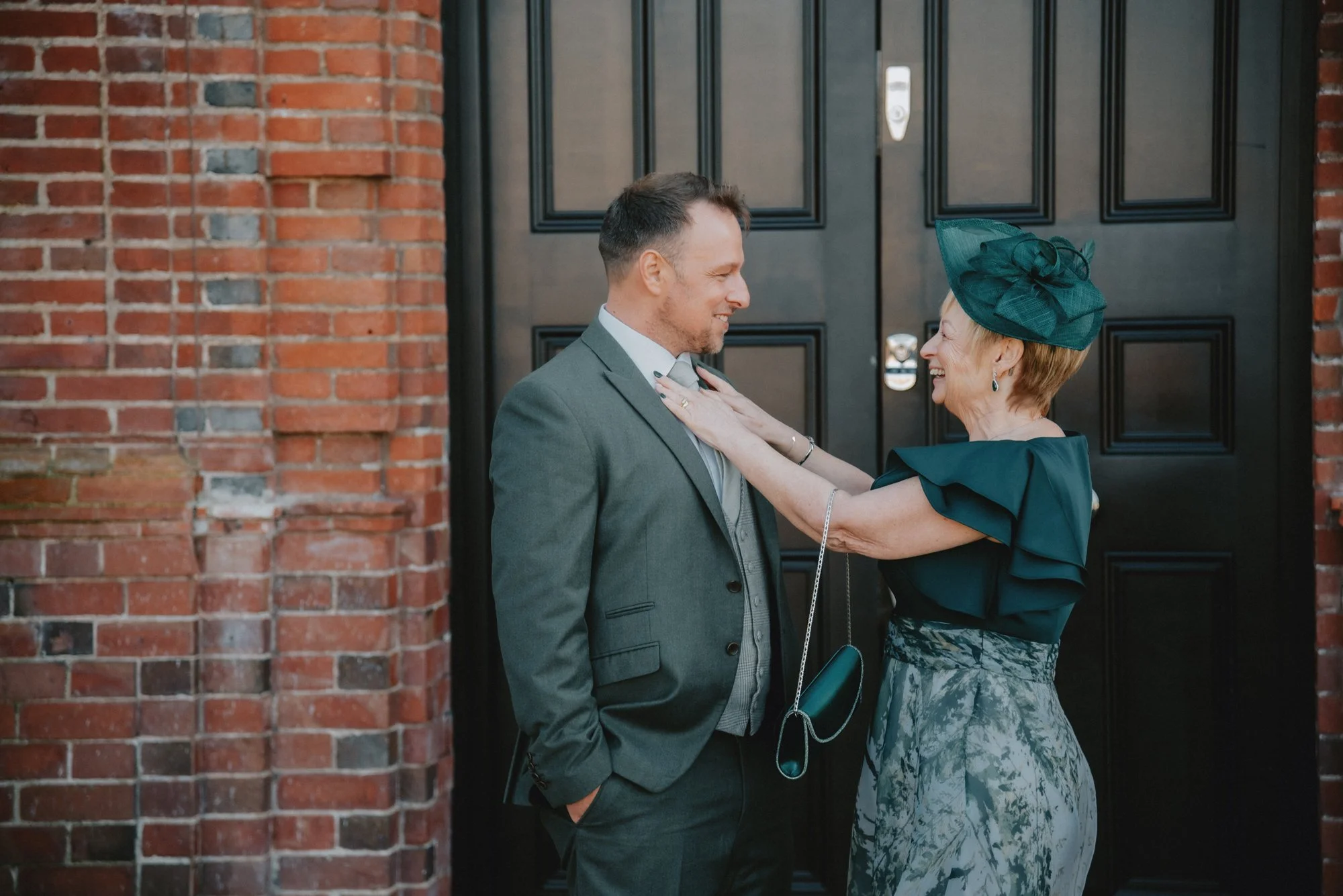 A man and an elderly woman facing each other in front of a black door. The woman is adjusting the man's tie and both are smiling, indoors or at the entrance of a building.