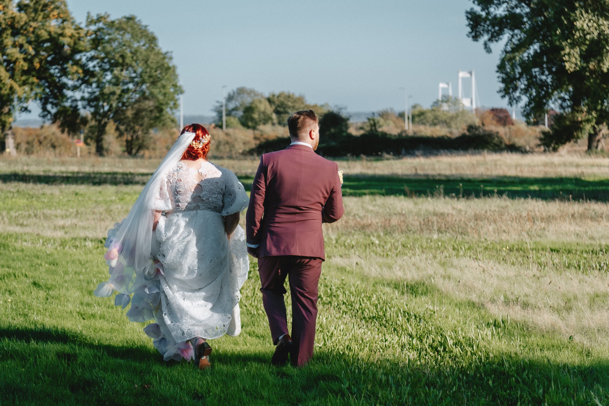 A couple in wedding attire walking on a grassy field with trees and a bridge in the background.