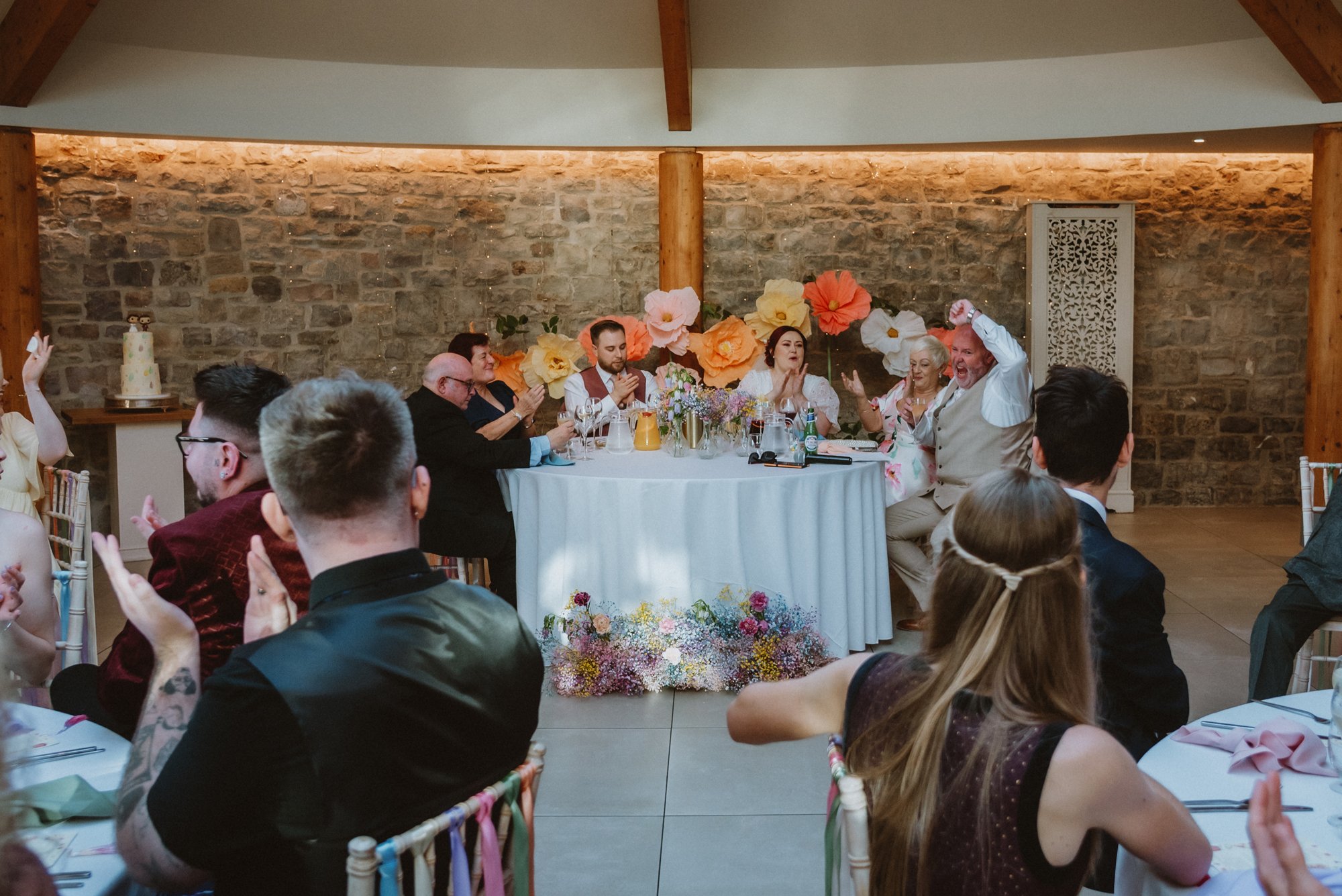 People seated at a wedding reception enjoying celebration, with a decorated table and colorful floral backdrop.