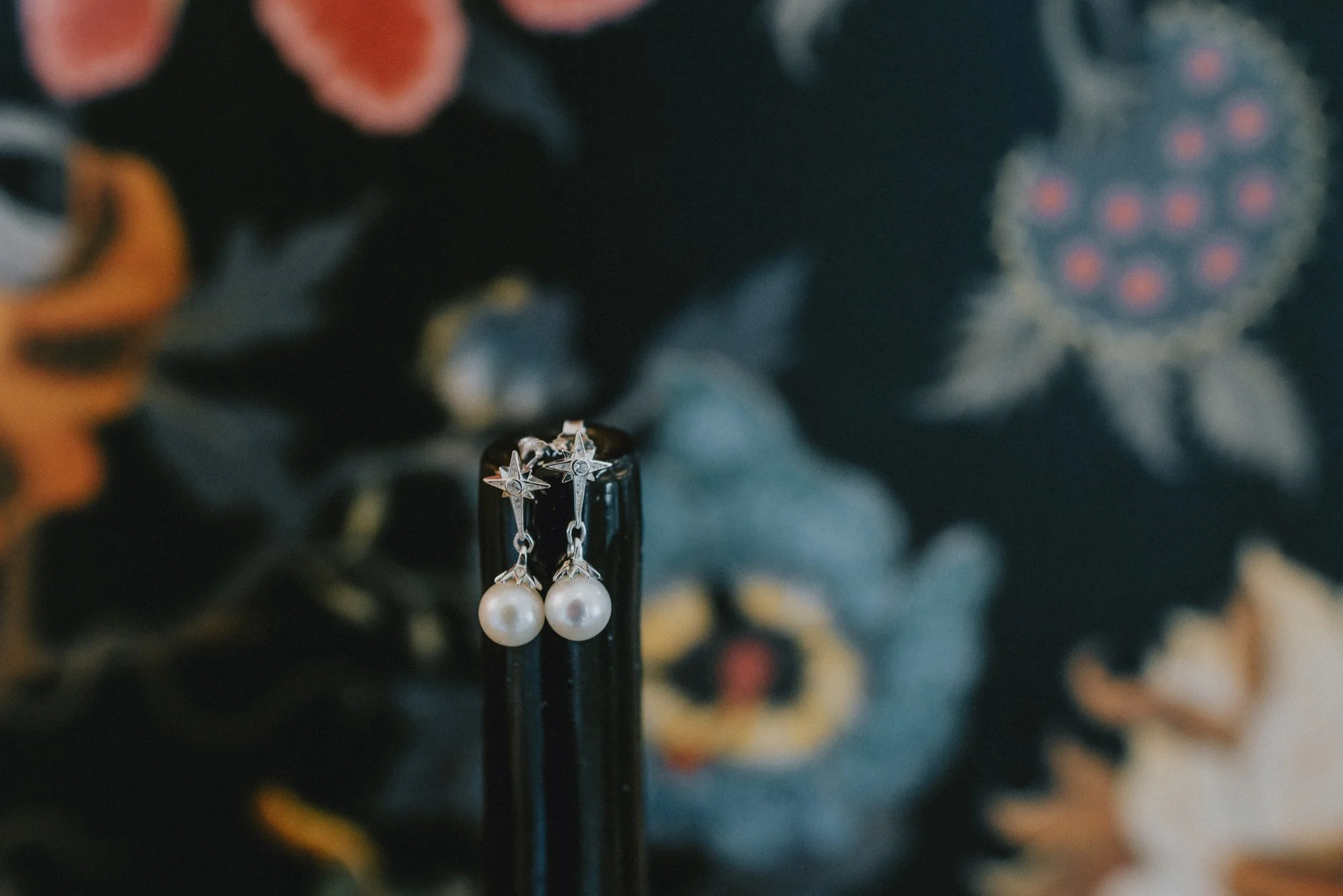 A pair of pearl drop earrings hanging from a black stand, with a blurred floral background.