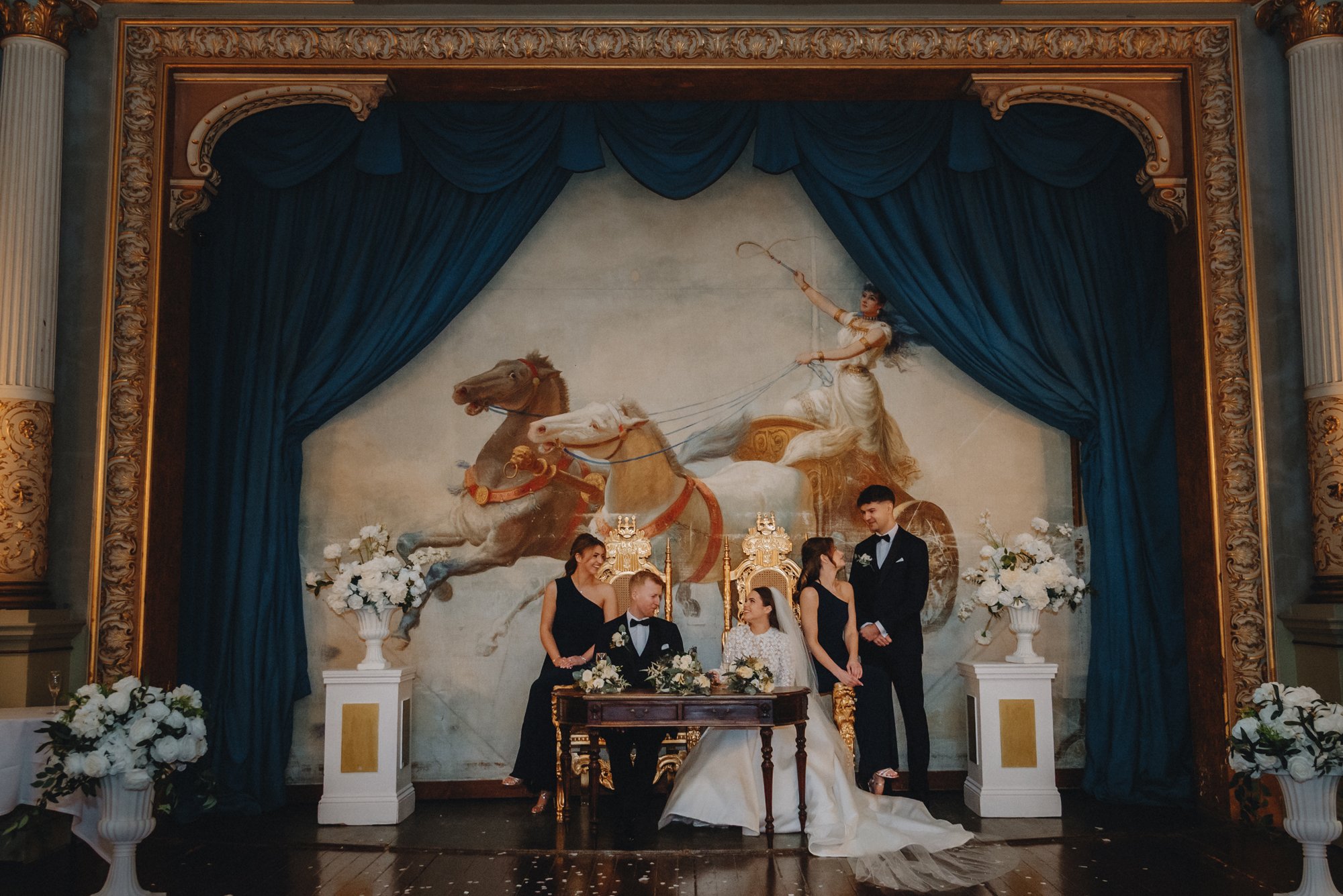 A wedding ceremony with a bride, groom, and four participants standing behind a table, with a mural of a chariot pulled by horses and a woman riding it, surrounded by curtains and floral arrangements.