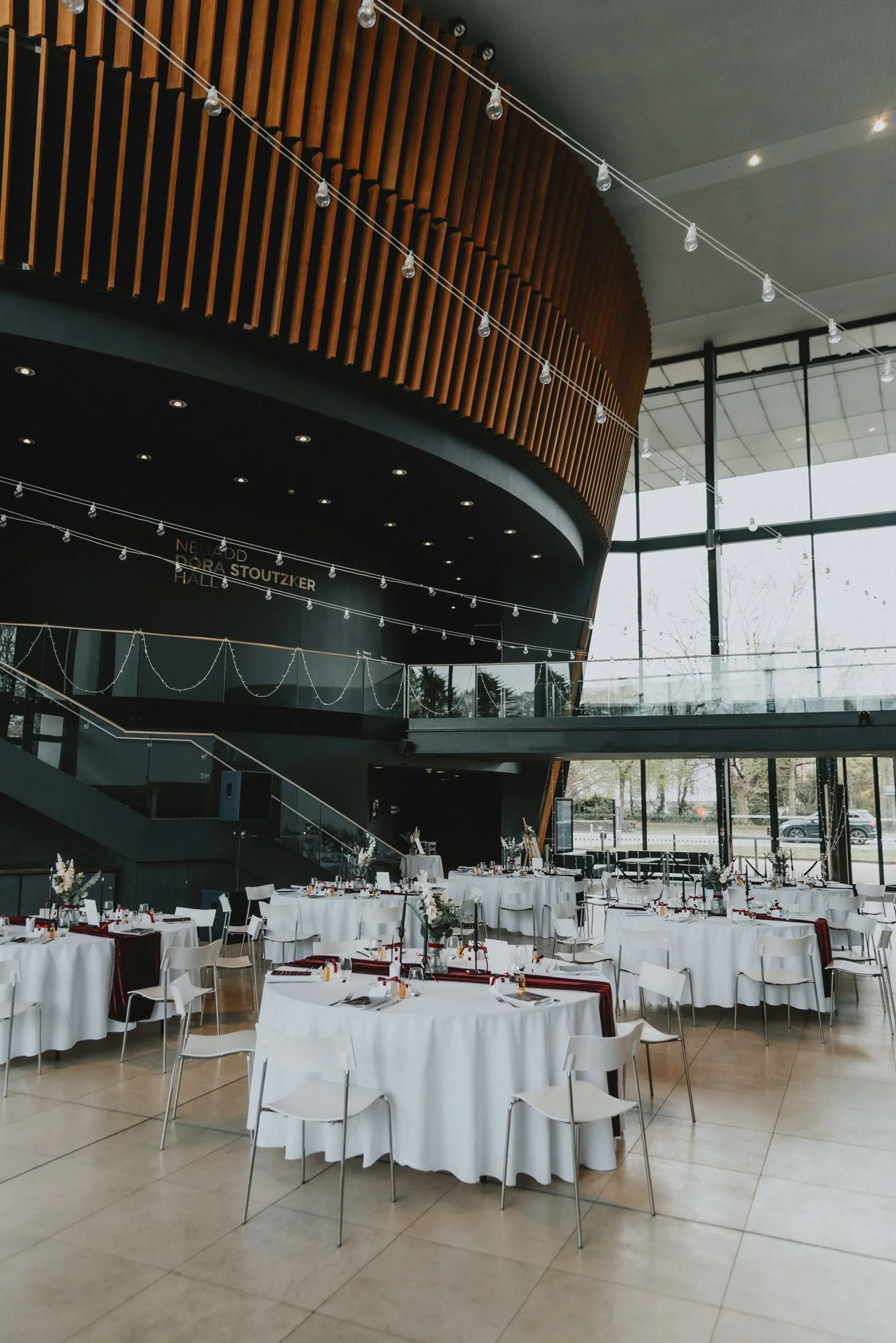 Inside a modern event hall with round tables covered in white tablecloths, set for a formal event, decorated with flower centerpieces and tableware. The hall features large floor-to-ceiling windows, a high ceiling, and a wall with wood paneling and s