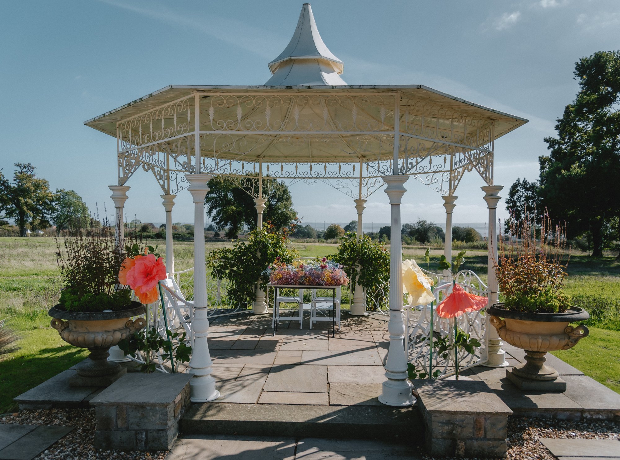 White ornate gazebo with flower arrangements on its perimeter, situated on a stone platform in a grassy field, with trees and blue sky in the background.