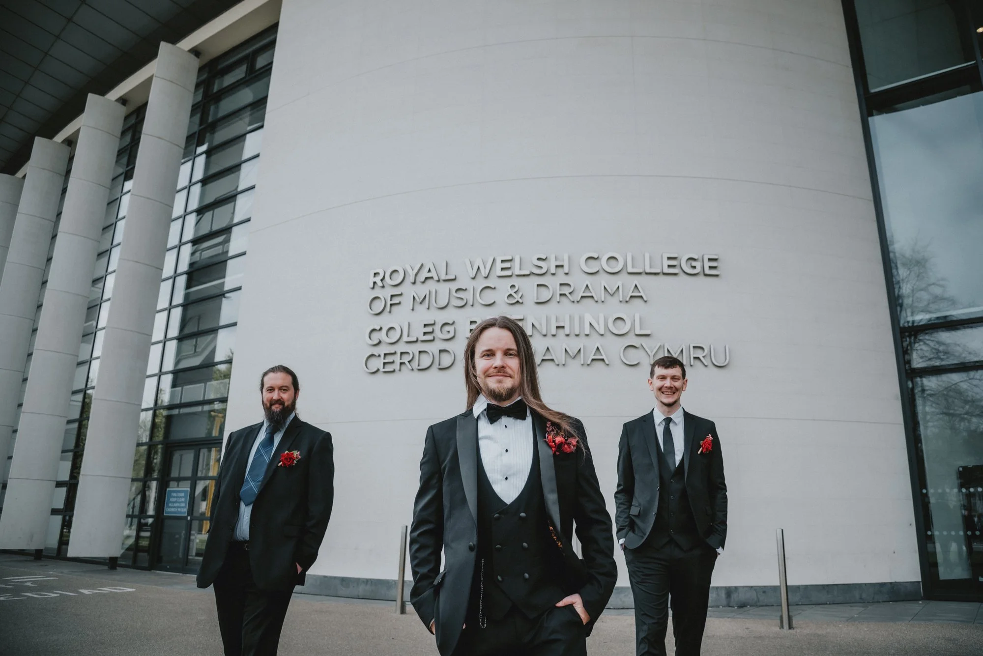 Three men in tuxedos standing outside the Royal Welsh College of Music and Drama, smiling, with the college's name on the building behind them.