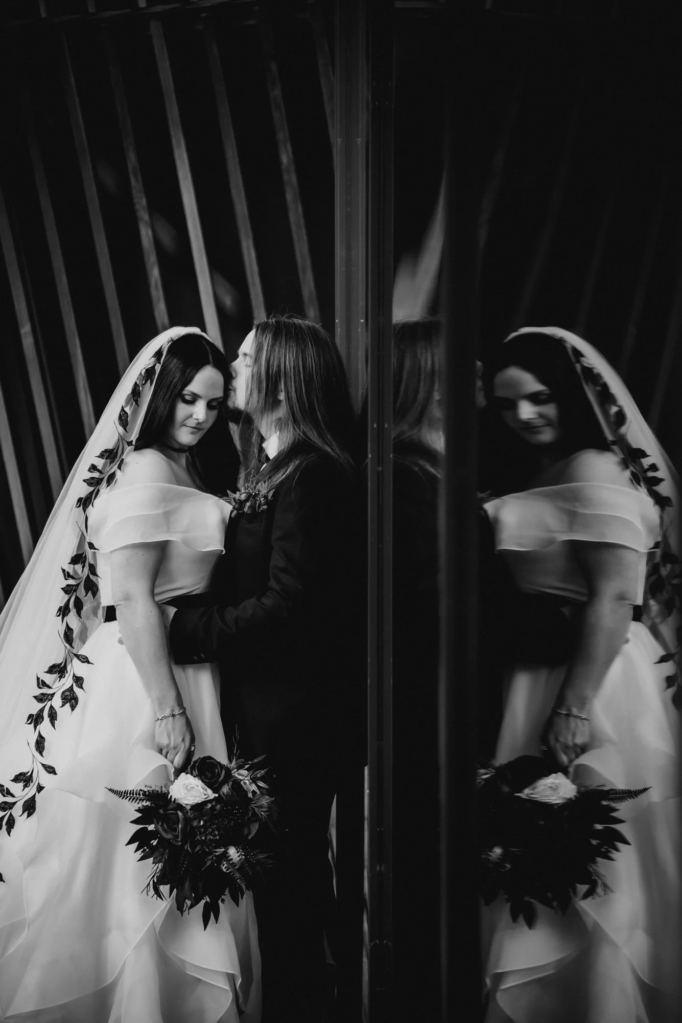 Black and white photo of a bride and groom standing close, with the groom kissing the bride's forehead. The bride is holding a bouquet and wearing a wedding dress and veil. The couple is reflected in a mirror, creating a symmetrical image.