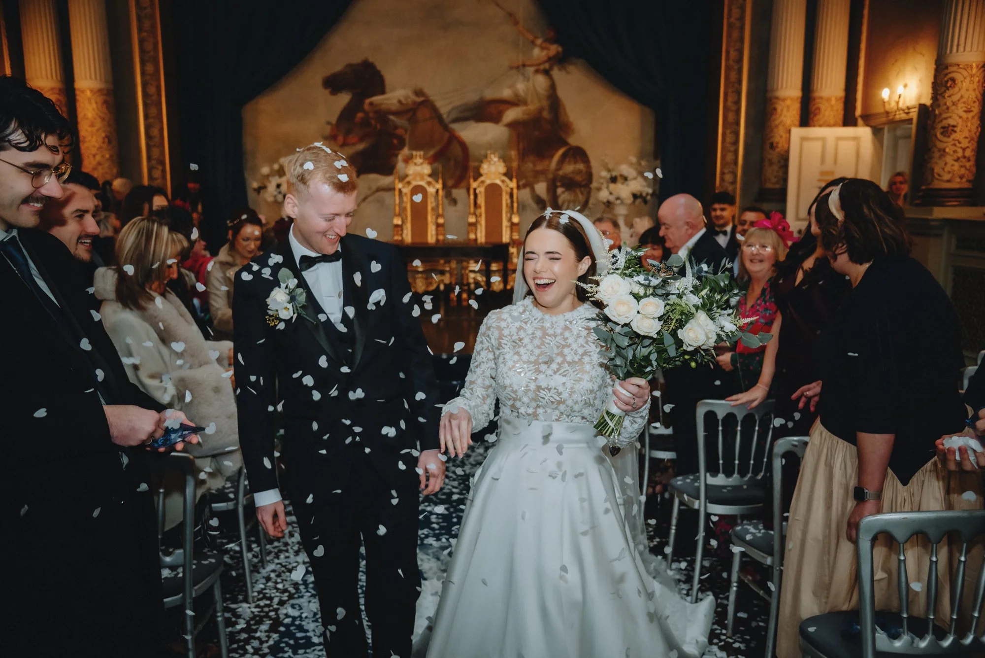 A bride and groom walking down the aisle after their wedding ceremony, smiling and holding hands, with guests celebrating and throwing confetti in a decorated indoor venue.